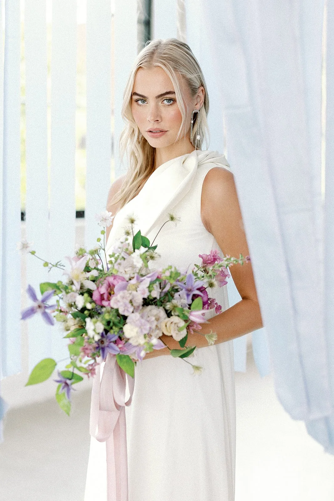 A young woman in a white dress holding a bouquet of pink, purple, and white flowers indoors.