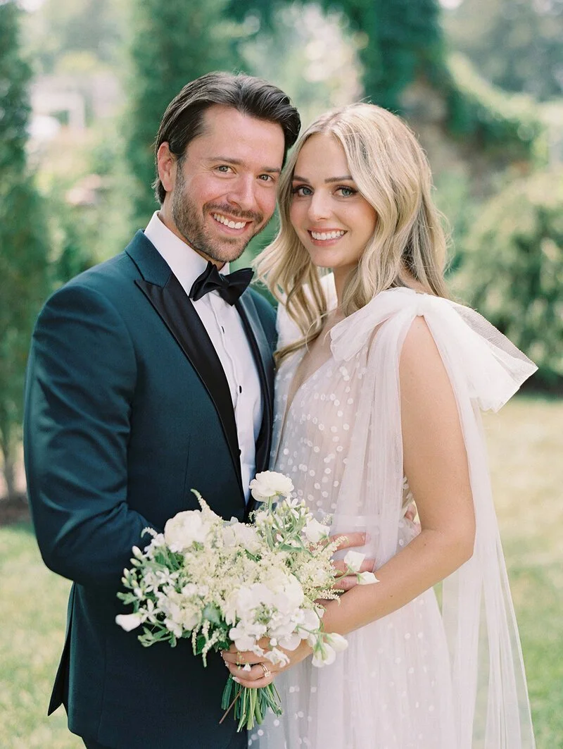 A happy couple on their wedding day, smiling outdoors with a green background. The groom wears a tuxedo with a bowtie, and the bride wears a white dress with tulle and holds a bouquet of white flowers.