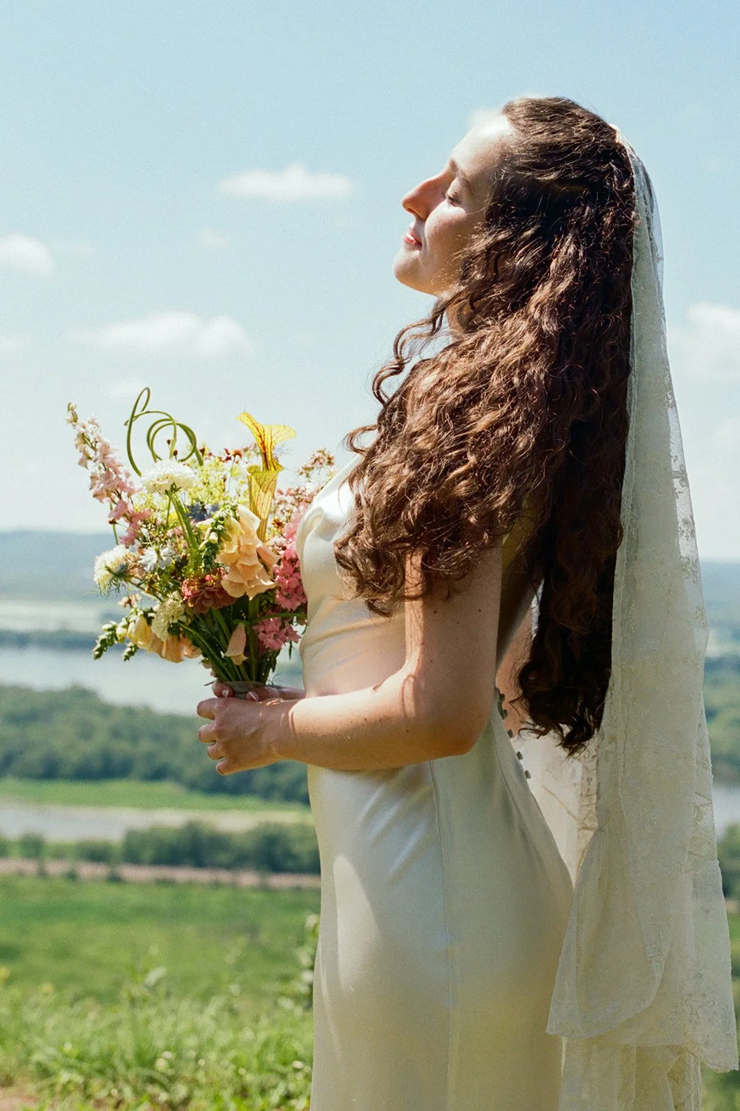 A woman on her wedding day, holding a bouquet of colorful flowers, with her eyes closed and face turned towards the sky, outdoors against a blue sky with clouds.