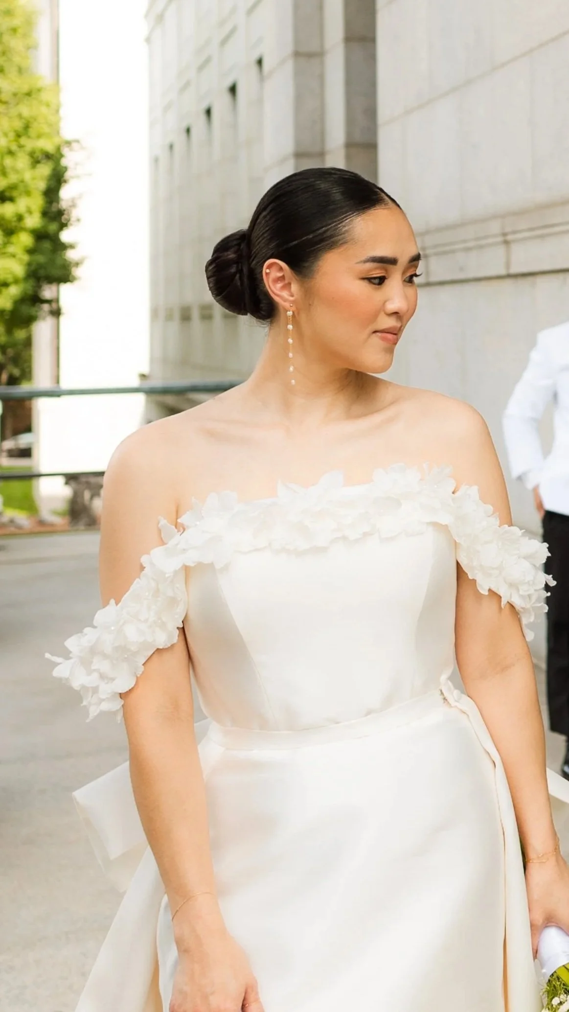 A woman in a white wedding dress with floral details on the shoulders, standing outdoors near a stone building, with her hair styled in a sleek bun and wearing drop earrings, looking to her right.