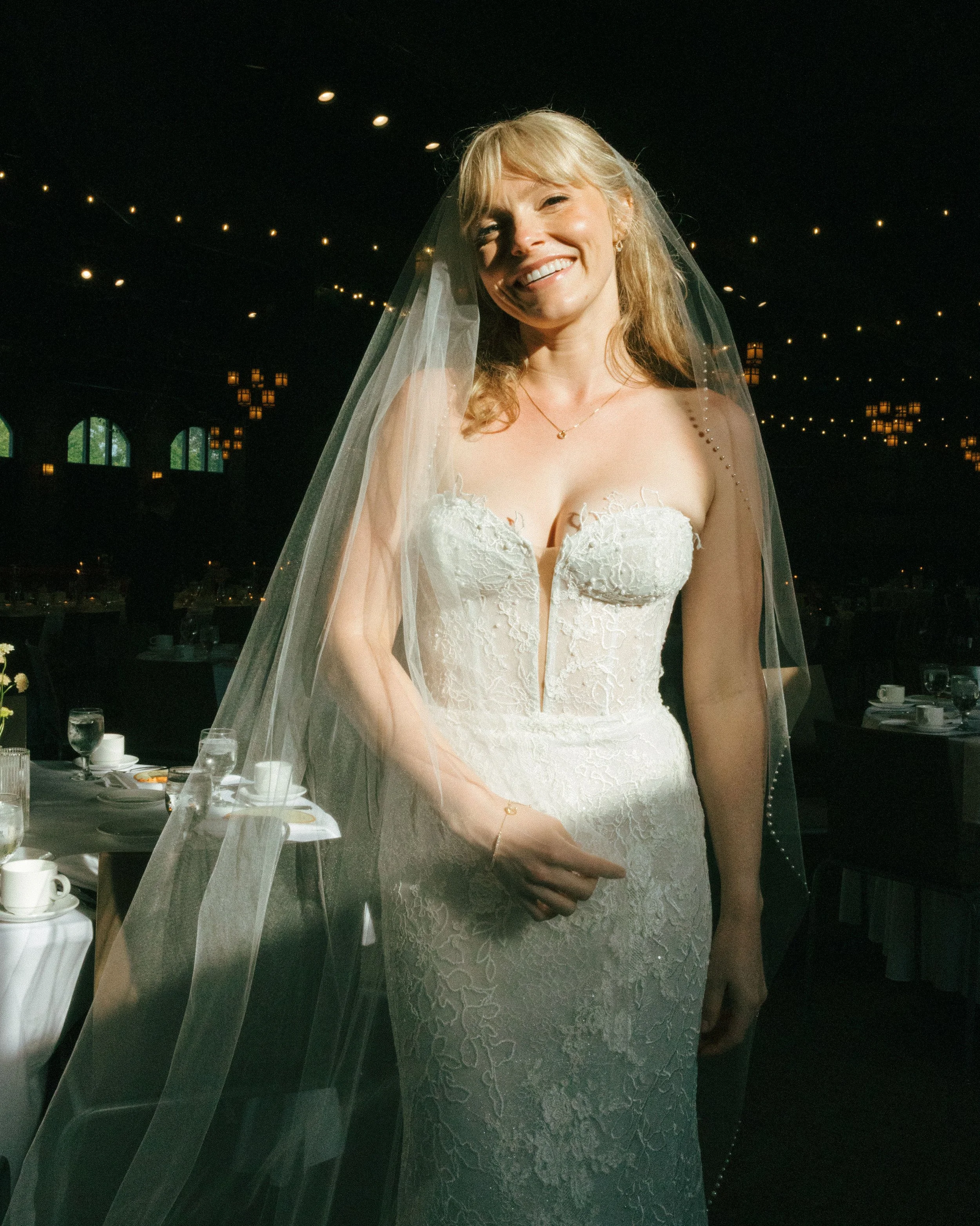 A smiling bride in a strapless lace wedding dress with a veil, standing inside a dimly lit venue with string lights and tables set for a celebration.
