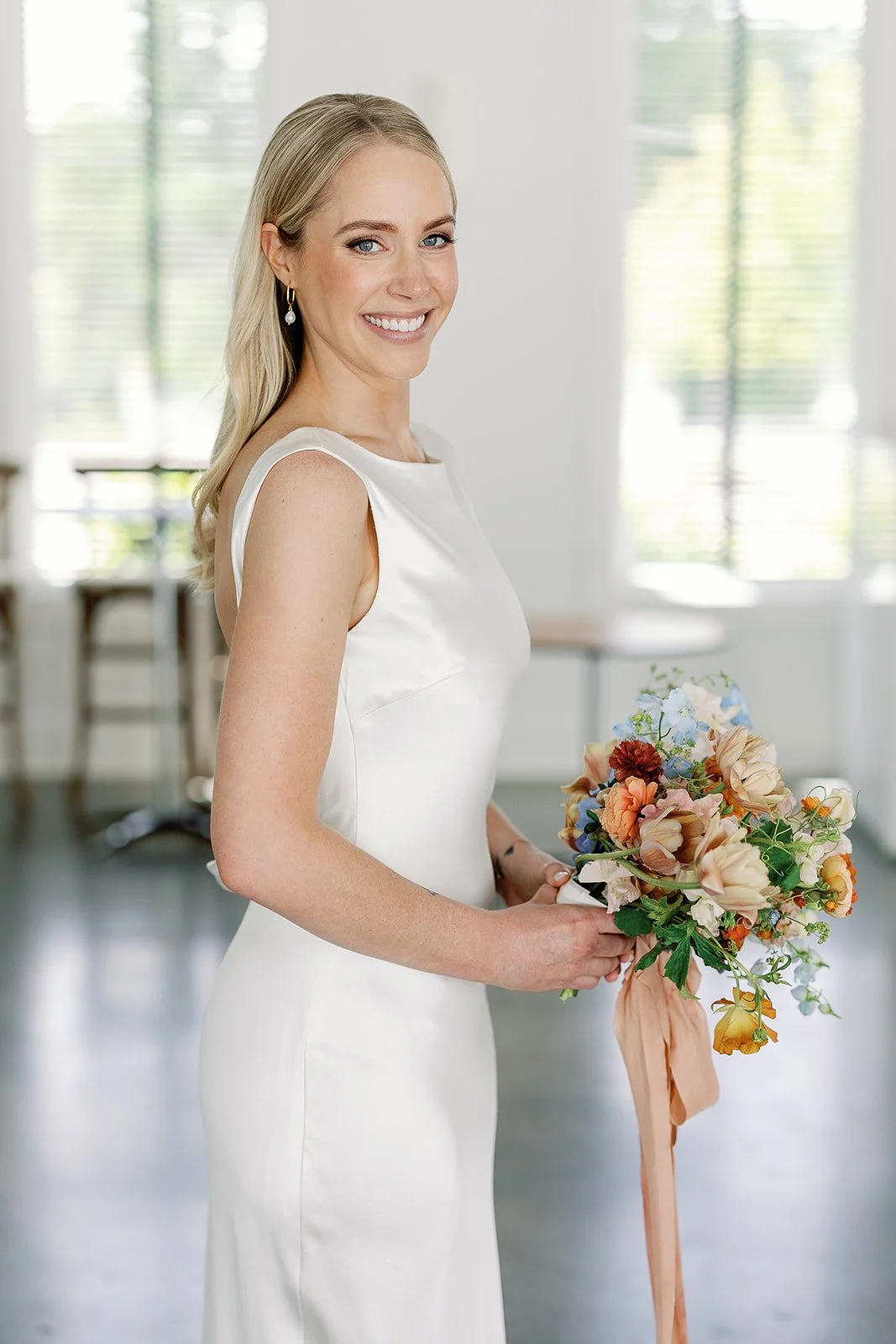 A smiling woman in a white dress holding a colorful bouquet of flowers in an indoor setting with large windows.
