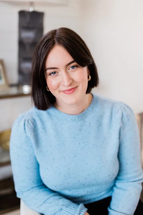 A woman with shoulder-length brown hair, wearing a light blue sweater and gold hoop earrings, smiling in an indoor setting.