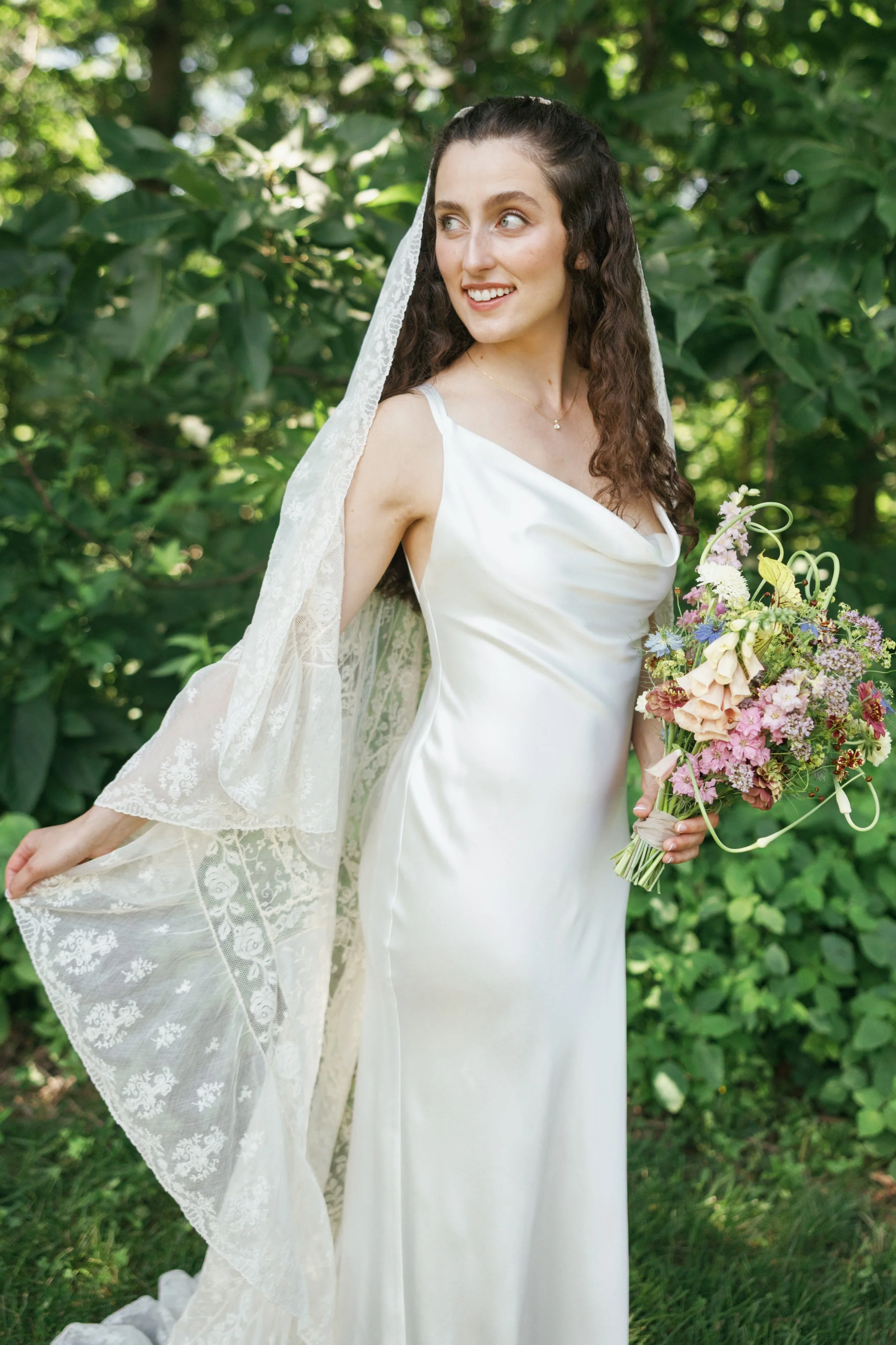 A young woman in a white wedding dress outdoors, holding a bouquet of flowers, wearing a lace veil, with greenery in the background.