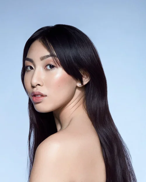Portrait of a young woman with long, dark hair and light makeup, looking at the camera against a light blue background.
