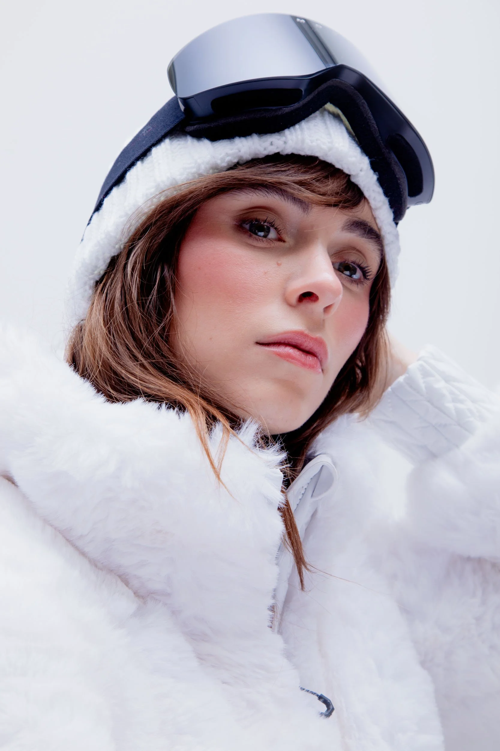 A close-up of a woman wearing ski goggles, a white knit hat, and a white fluffy jacket, outdoors in a snowy environment.