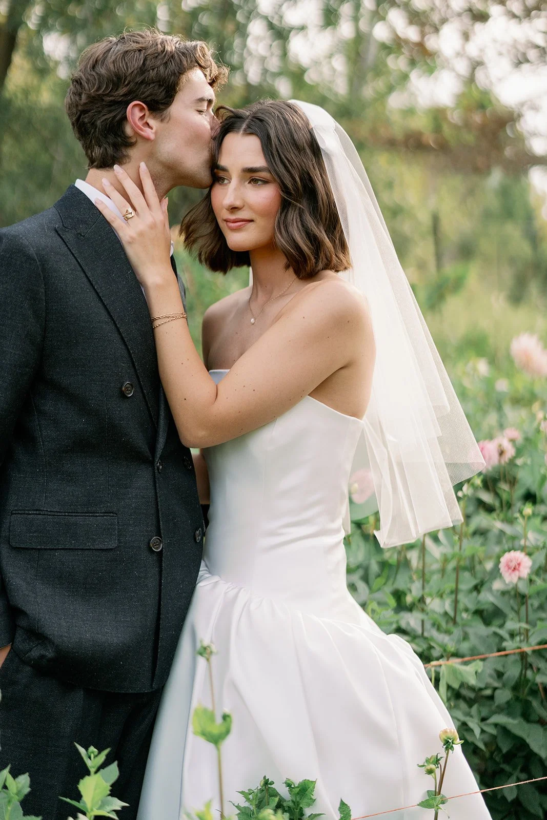 A bride in a white strapless wedding dress and a groom in a dark suit stand close together outdoors, with the groom kissing the bride's forehead. The bride has shoulder-length dark hair, wears a veil, and looks content. The background is green with pink flowers.
