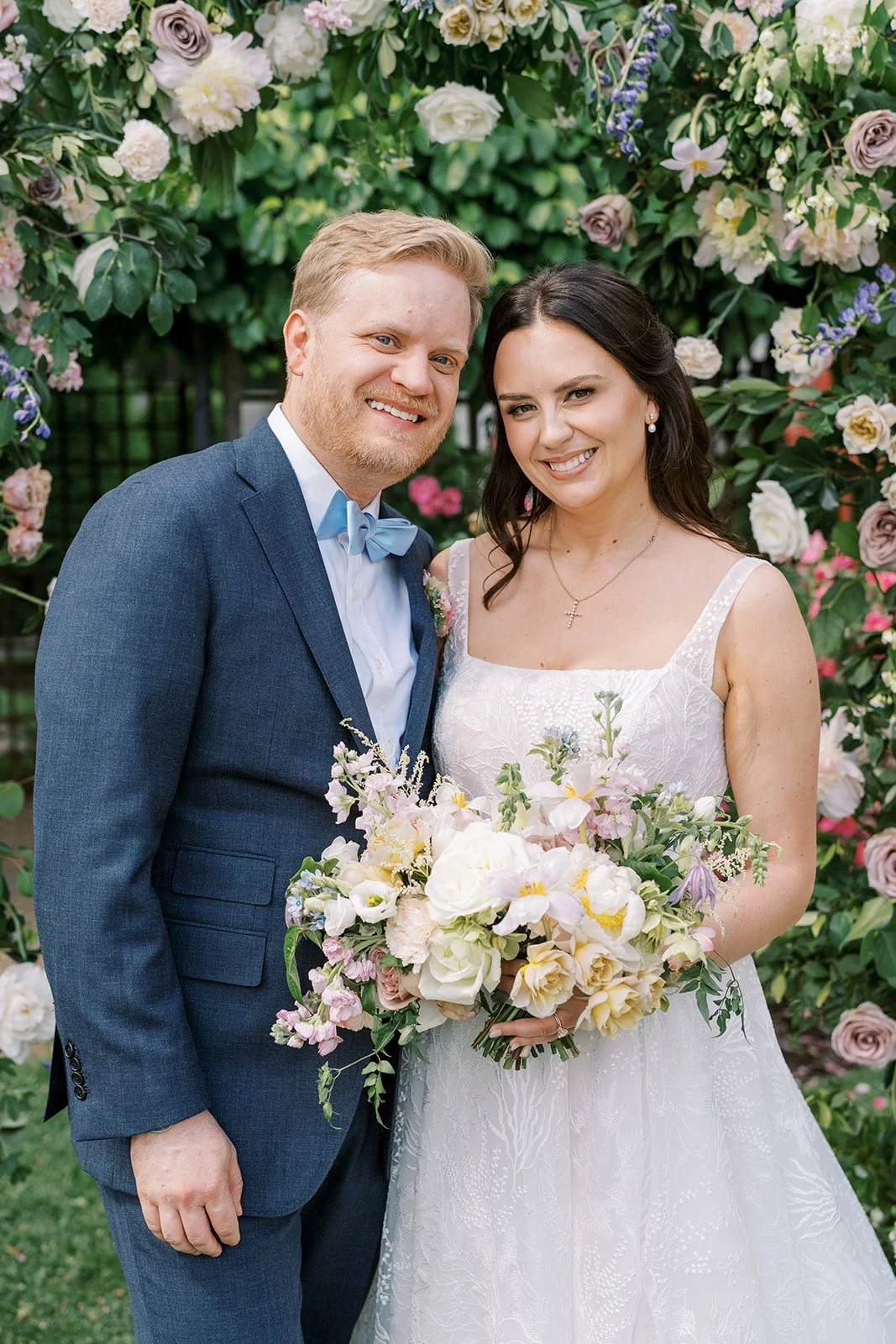 A smiling bride and groom standing in front of a lush flower backdrop during their wedding. The bride holds a large bouquet of white and pastel flowers, and the groom wears a blue suit with a light blue bow tie.