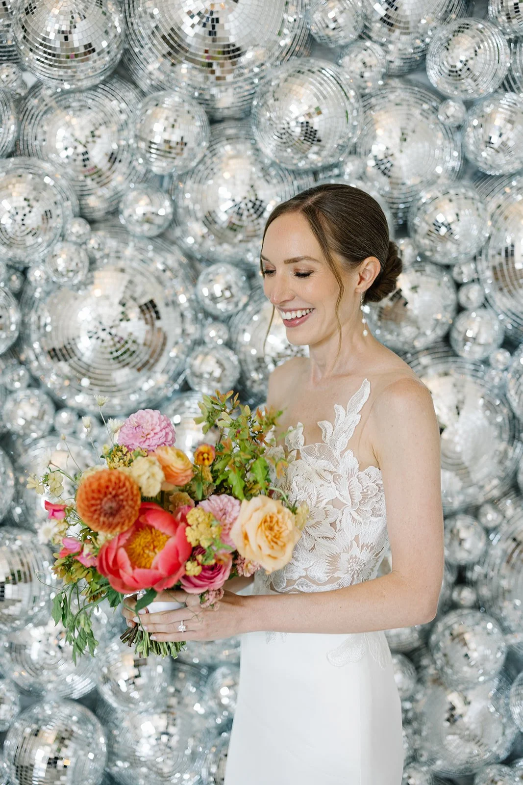 A bride smiling while holding a colorful bouquet of flowers in front of a background filled with shiny, reflective disco balls.