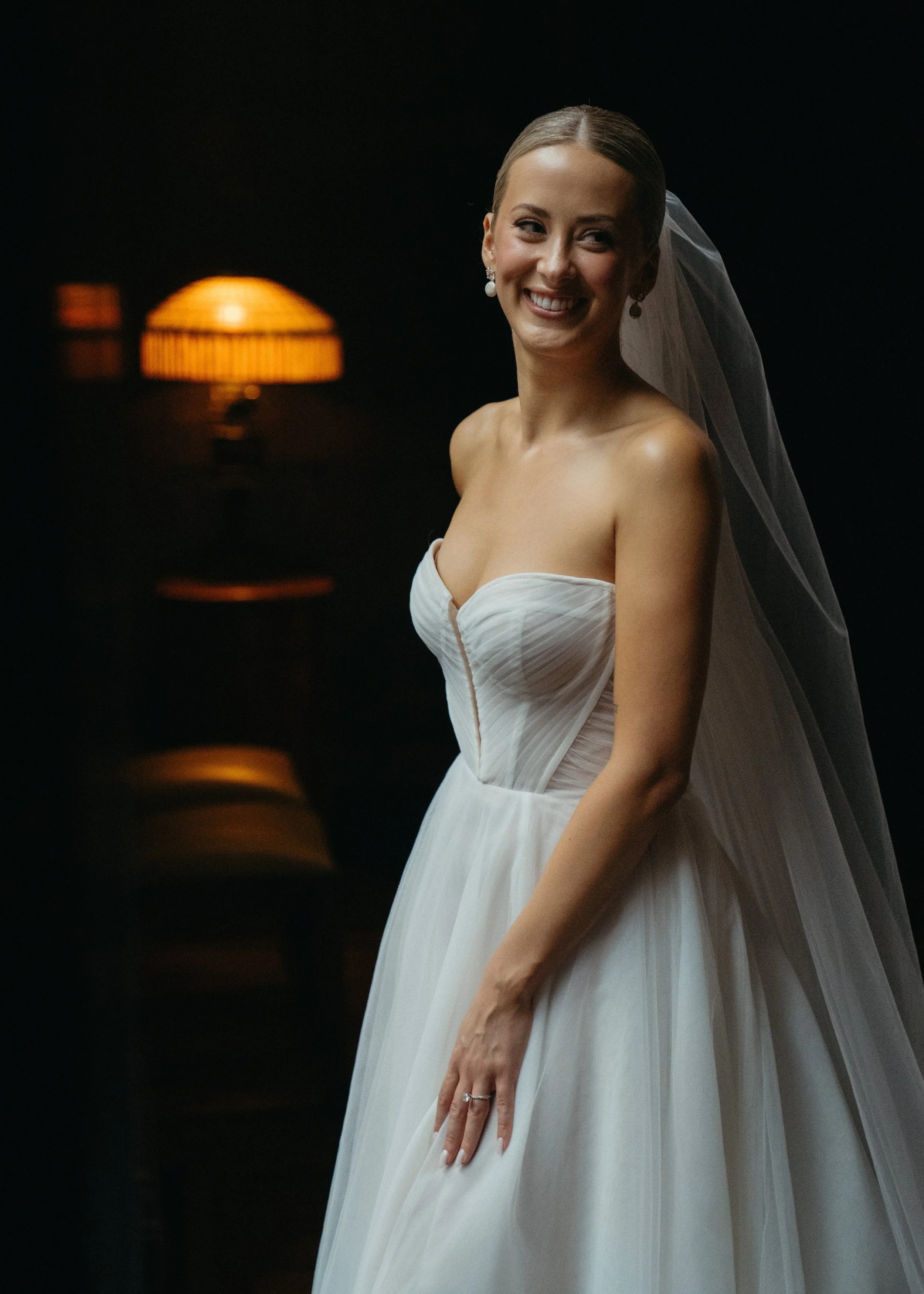 A smiling bride with jewelry and a veil, wearing a strapless white wedding gown, standing against a black background with warm-toned lighting.