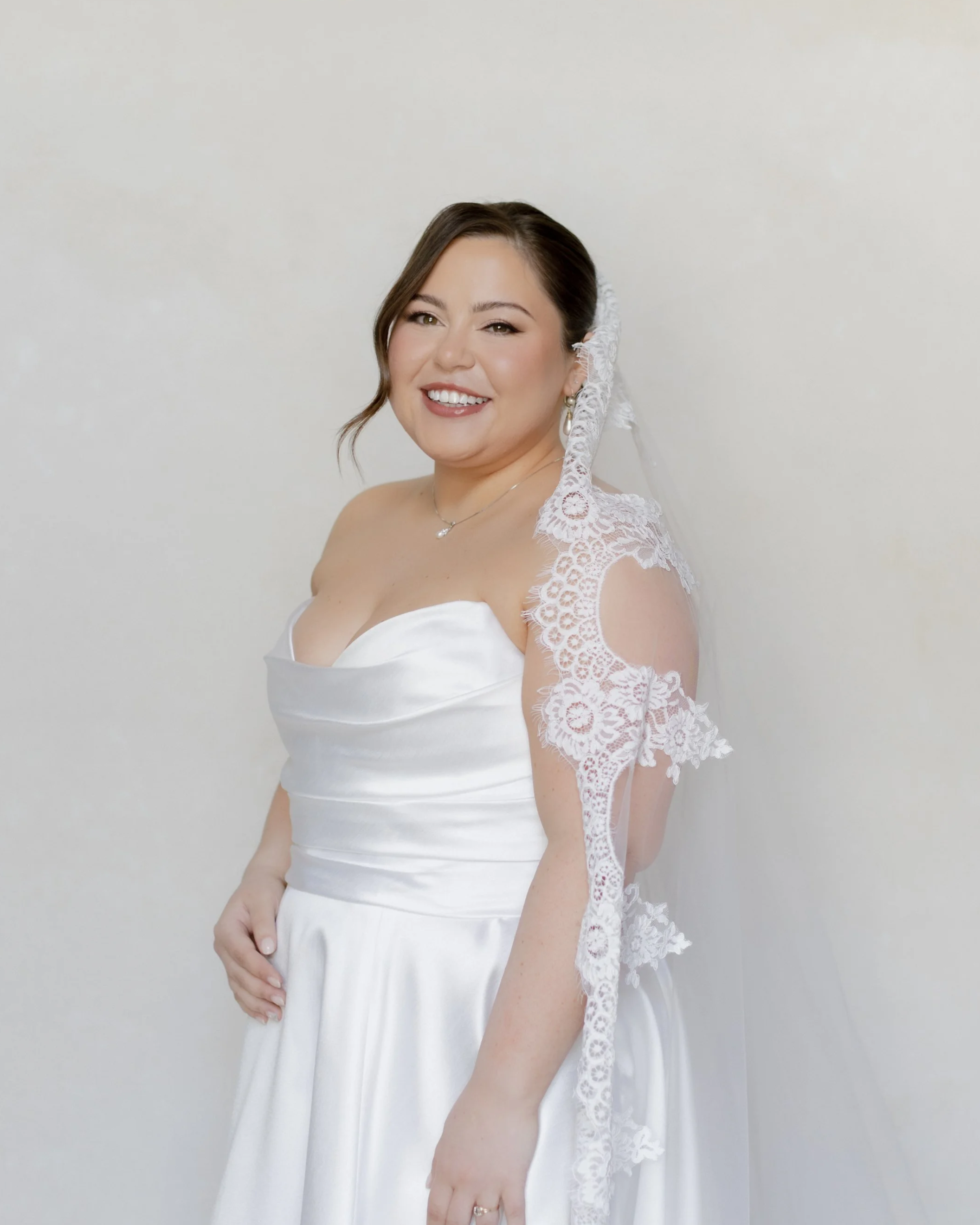 A smiling woman in a white lace wedding dress with off-the-shoulder lace sleeves, standing in front of a plain light background.