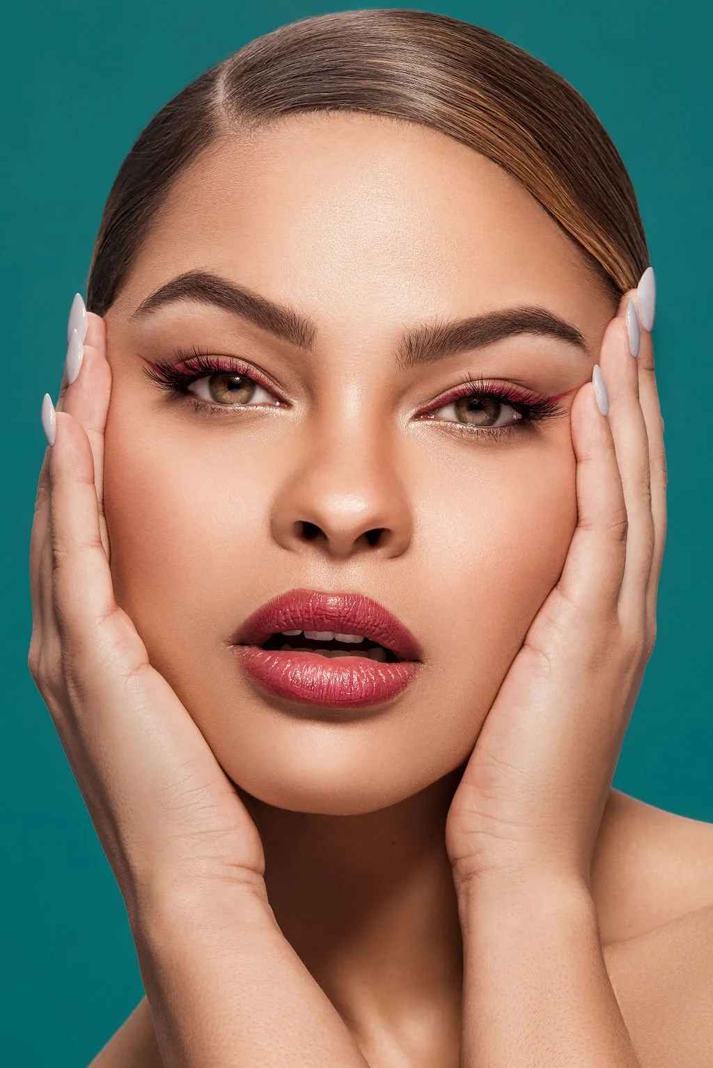 Close-up of a woman with flawless skin, wearing makeup including pink eyeshadow, long lashes, and red lipstick, holding her face with both hands, against a teal background.