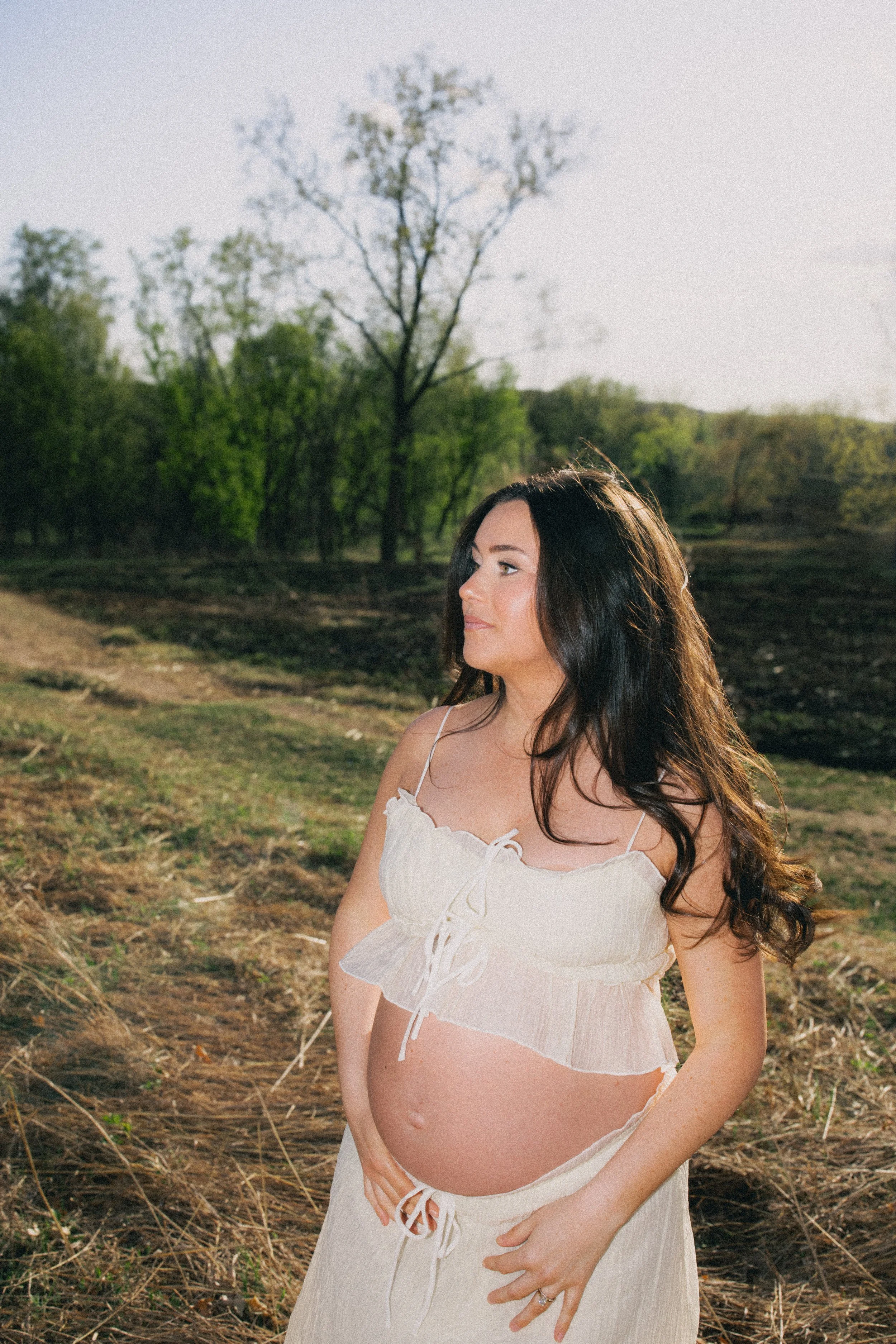 A pregnant woman with long dark hair standing outdoors in a field of grass and trees, wearing a white sleeveless dress with ruffles and a bow tied at her waist, looking to her right with a serene expression.