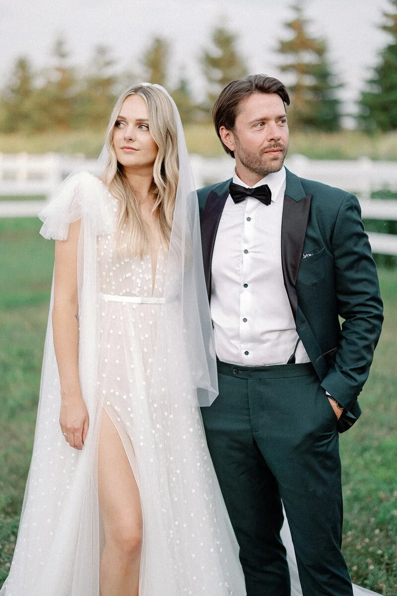 A bride in a white wedding dress with a slit, standing next to a groom in a dark suit with a bow tie. They are outdoors near a white fence with trees in the background.