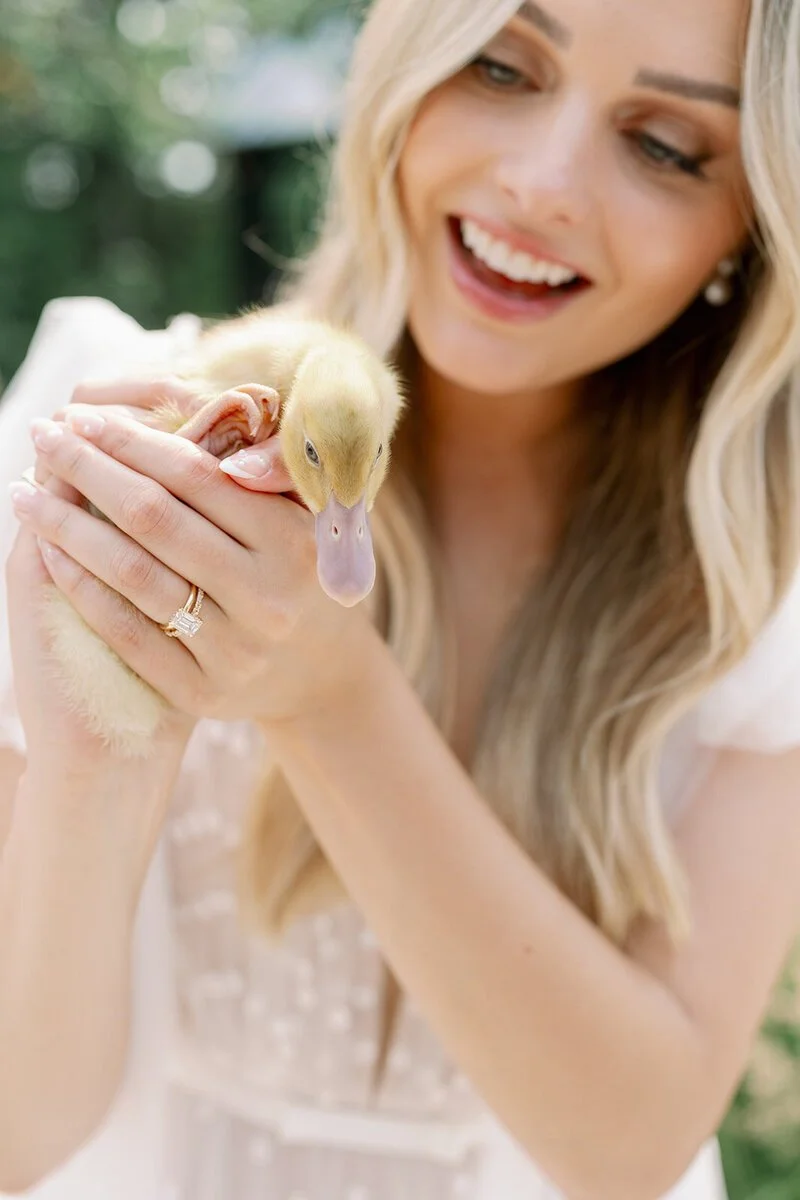 Woman holding a yellow duckling with greenery in the background.