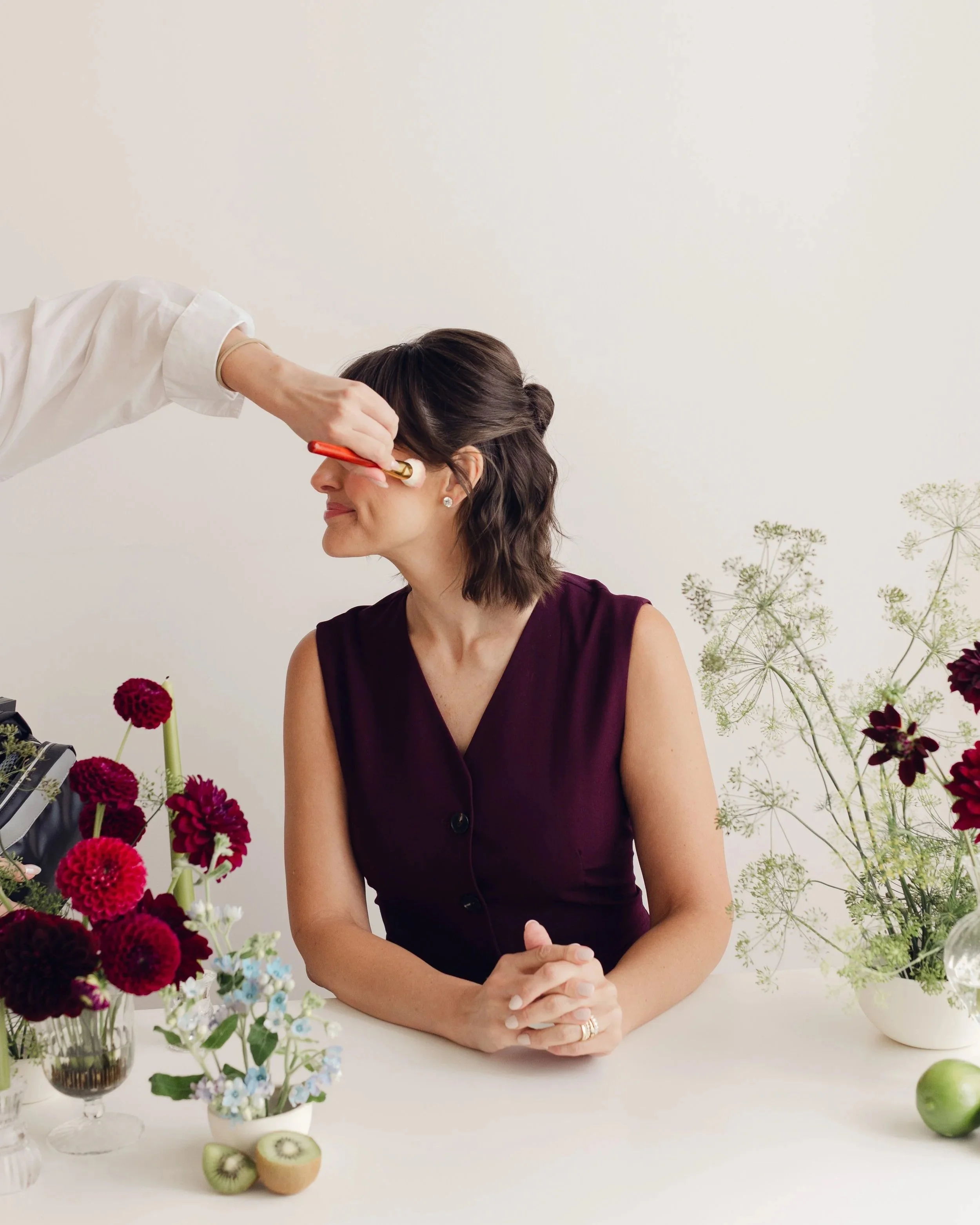 A woman with brown hair sitting at a table with flowers and fruit, having makeup applied by another person wearing a white coat. Photo Credit: Julia Mary Photography