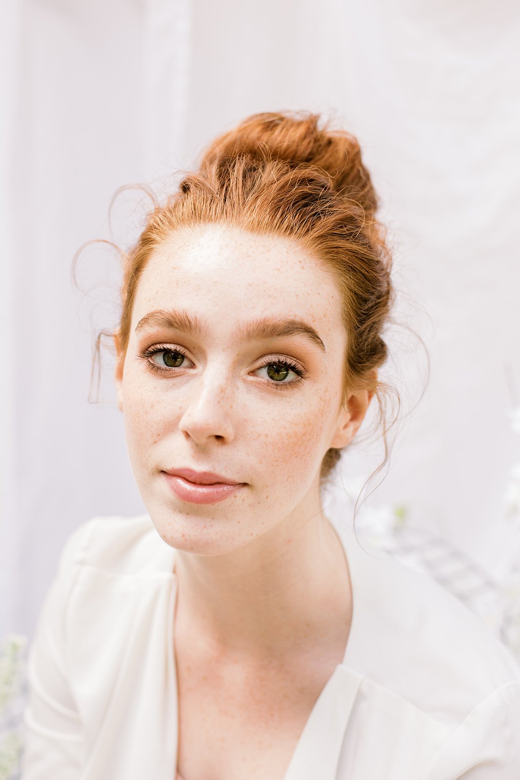 Close-up of a woman with red hair styled in an updo, wearing a white top with a soft smile, set against a light, neutral background.