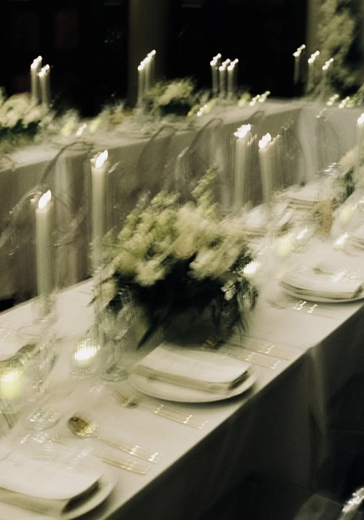 Elegantly set dining table with white linens, floral centerpiece, tall candles, and plates with napkins, in a dimly lit environment.