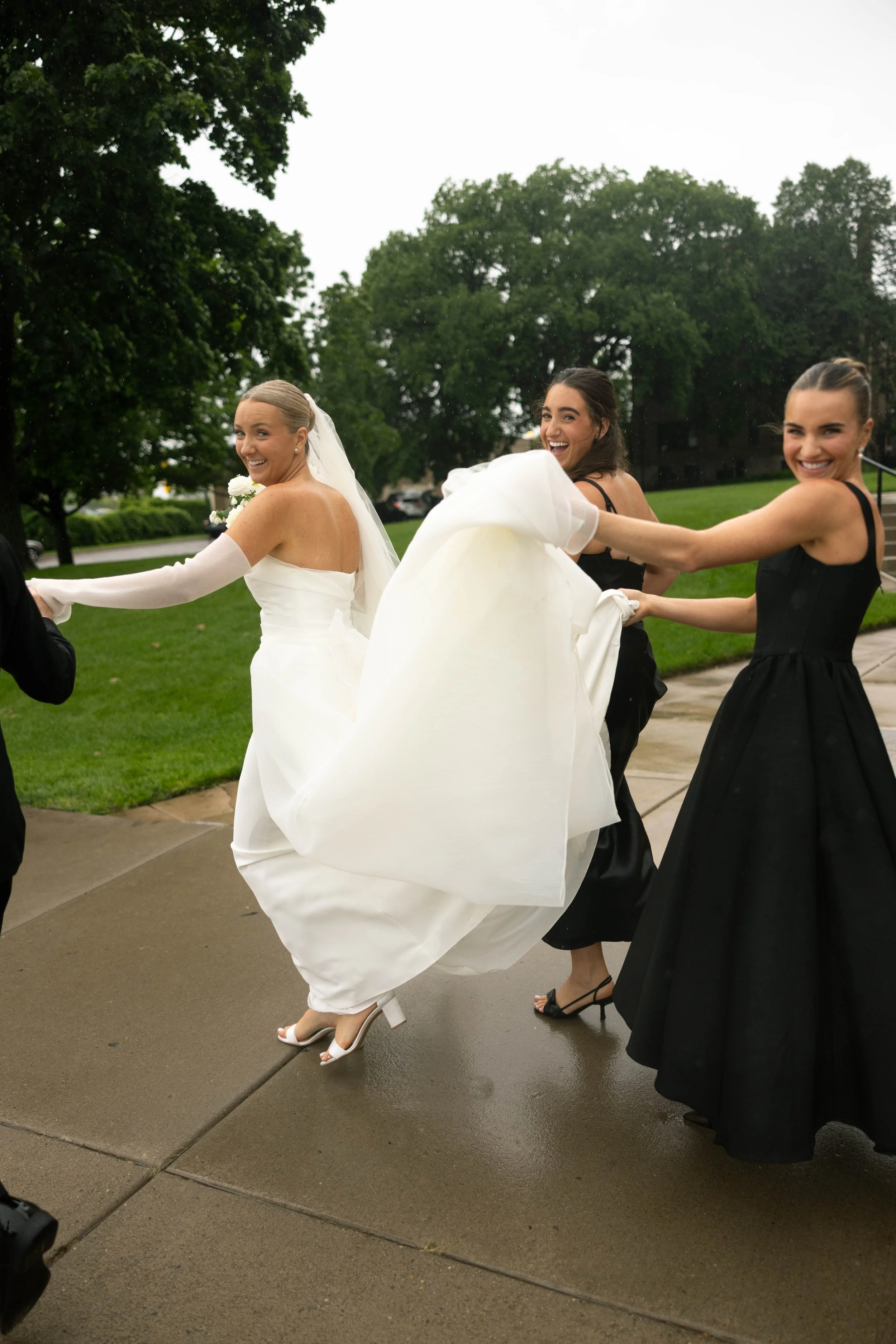 A bride and two bridesmaids playfully tug at the train of the bride's wedding gown outside on a wet sidewalk, smiling and laughing amid green trees and a cloudy sky.