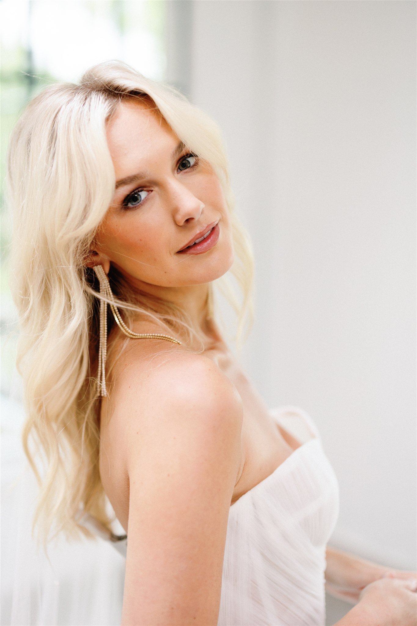 A woman with long blonde hair styled in loose waves, wearing dangling earrings, and dressed in a white off-shoulder top, standing in a bright room with natural light.