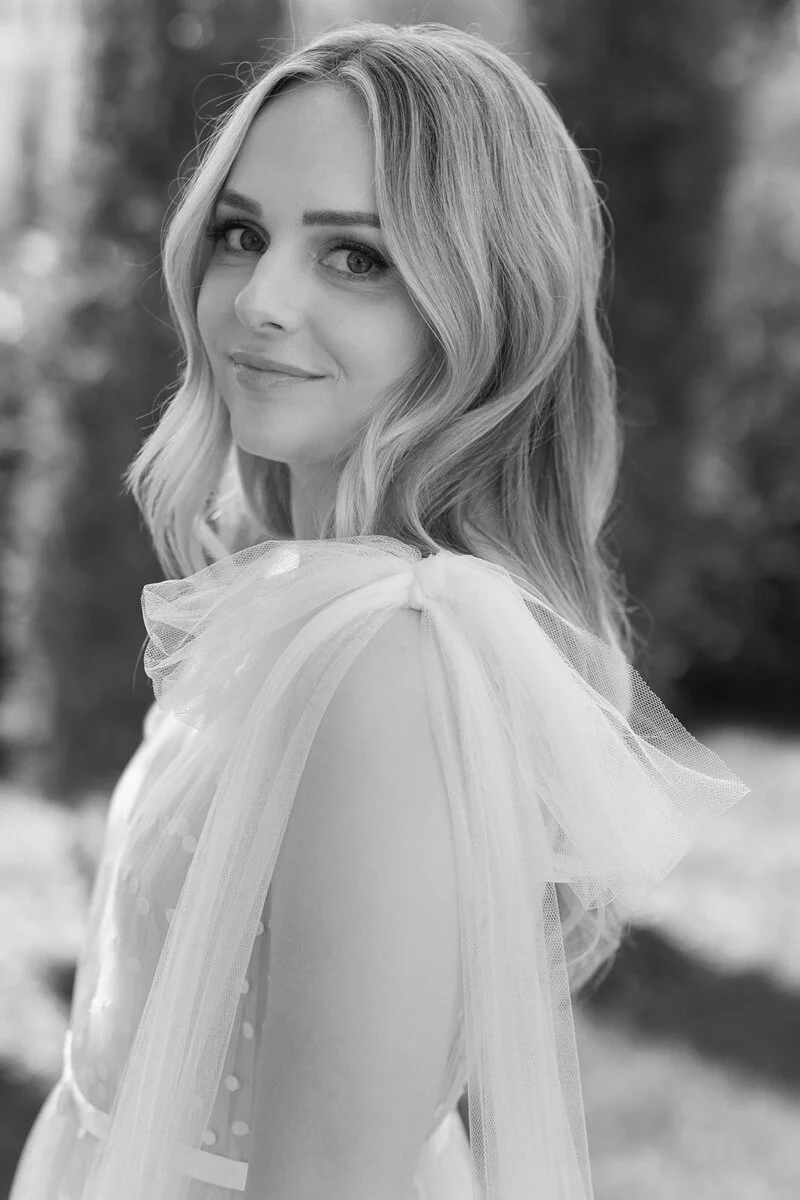 Black and white portrait of a young woman with wavy blonde hair, wearing a delicate dress with tulle details, outdoors.