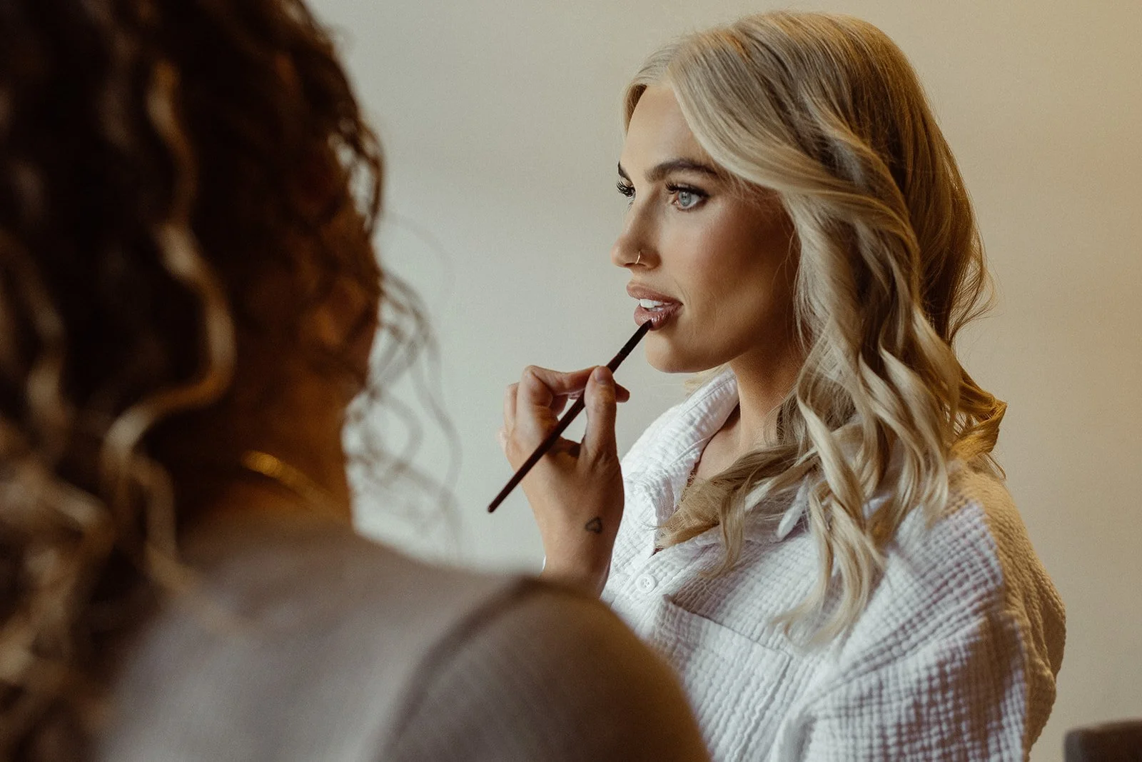 A woman with blonde hair getting her makeup done by another person, who is applying lipstick or lip gloss with a brush.