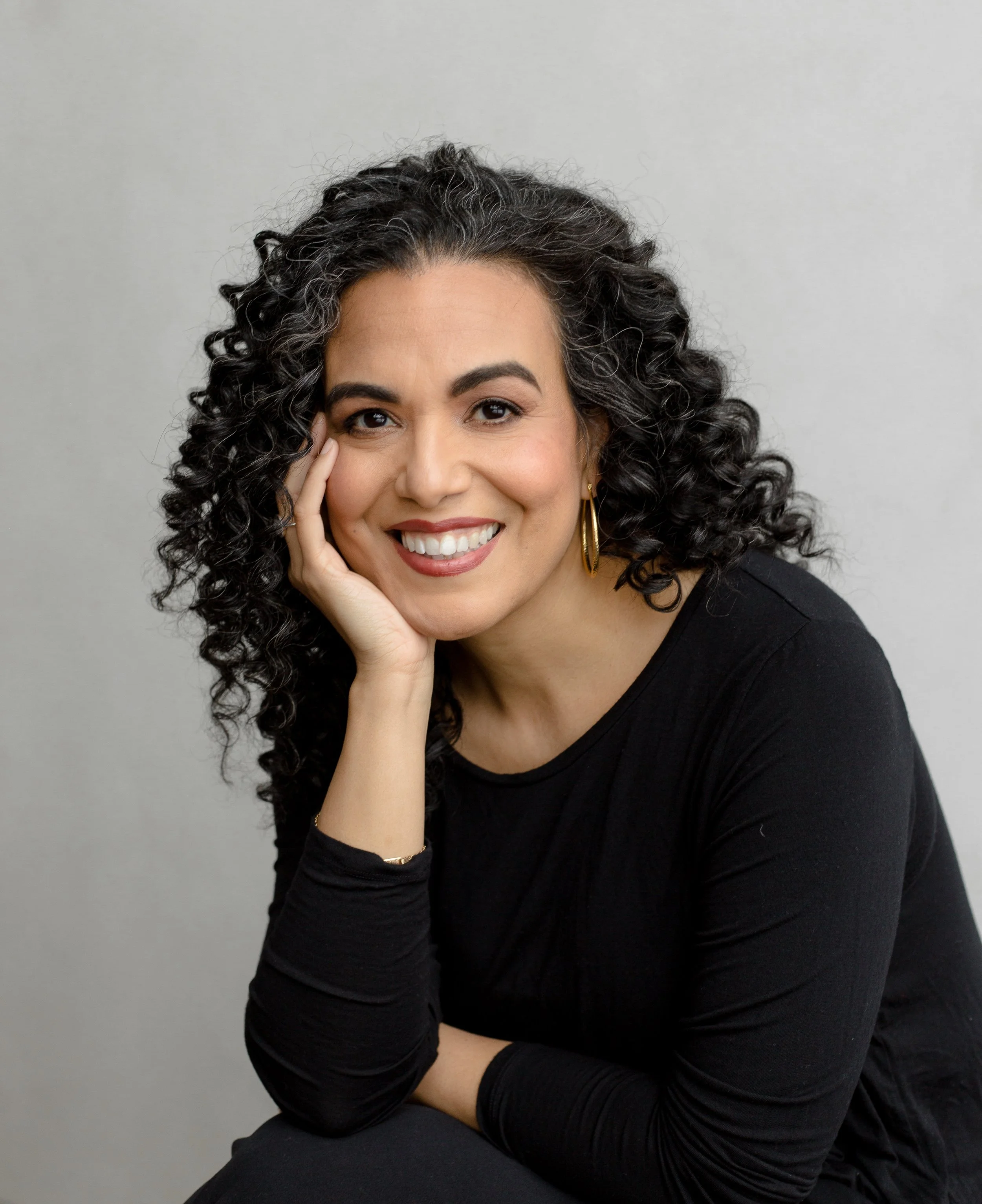 A woman with curly dark hair and wearing gold hoop earrings smiling with one hand resting on her face, dressed in a black top, against a plain gray background.