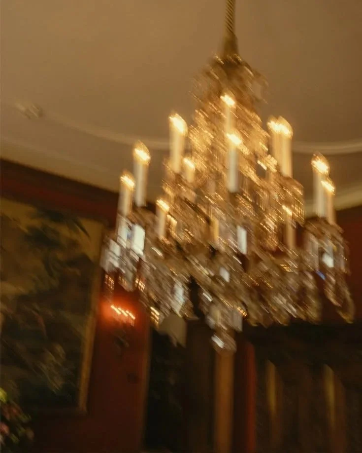 A large, ornate chandelier with several candle-shaped light bulbs hanging from the ceiling in a room with dark walls and a painting.