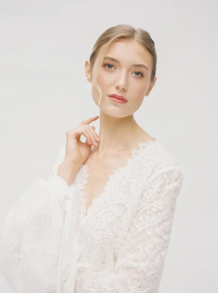A young woman with blonde hair and blue eyes wearing a white lace top, posing against a plain white background.