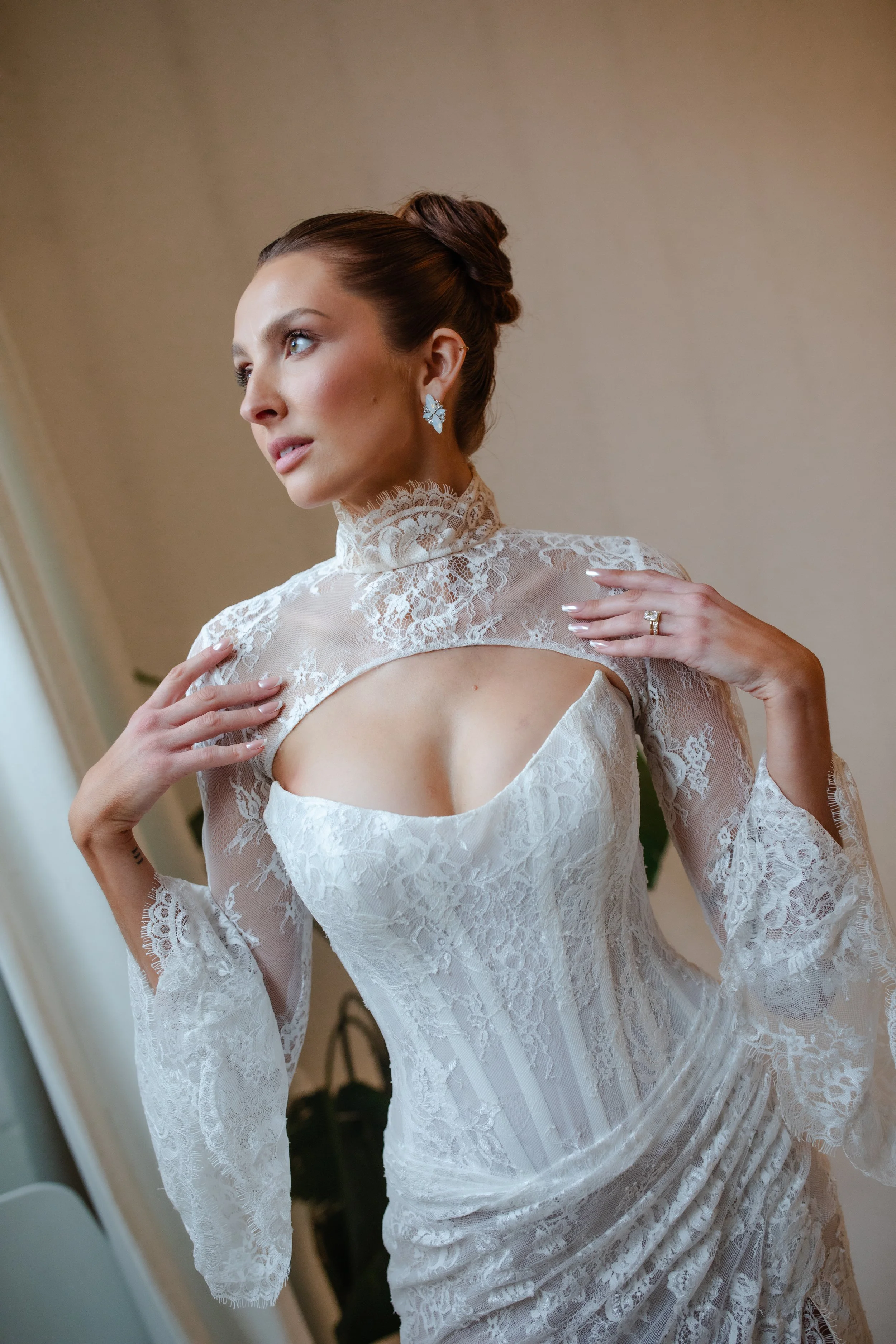 A woman with brown hair styled in an elegant updo, wearing lace earrings, a lace high-neck top, and a white dress with lace details and long, flowing sleeves. She is touching her shoulder and looking to the side.