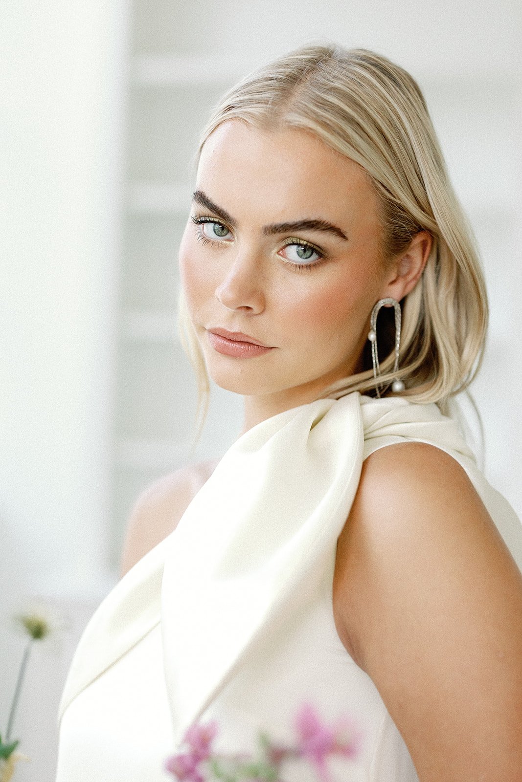 A young woman with blonde hair, blue eyes, wearing earrings and a white dress, looking at the camera in a bright, neutral background.