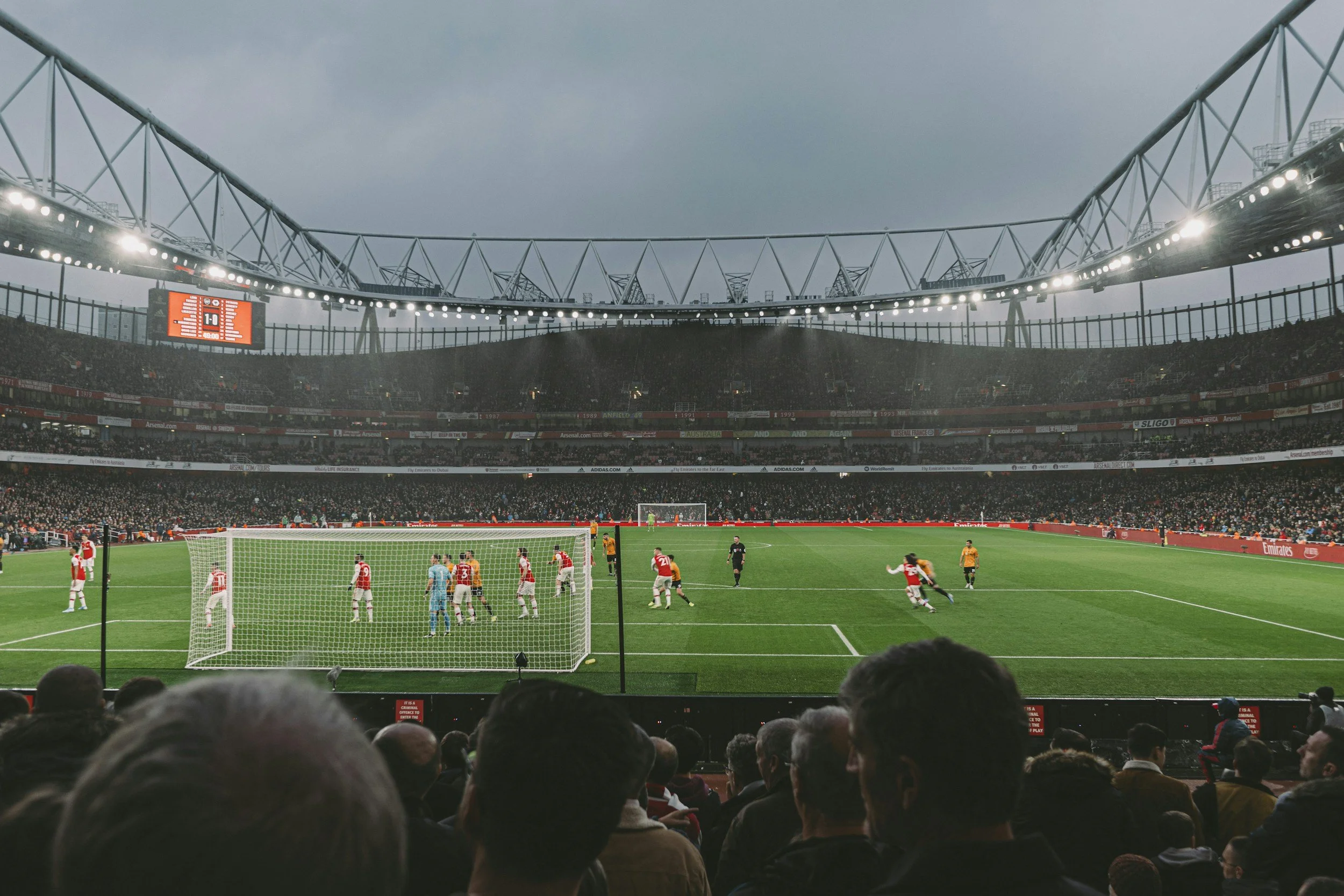 A stadium filled with spectators watching a soccer match. Players are on the field, with some near the goal. The stadium has bright lights and a cloudy sky overhead.