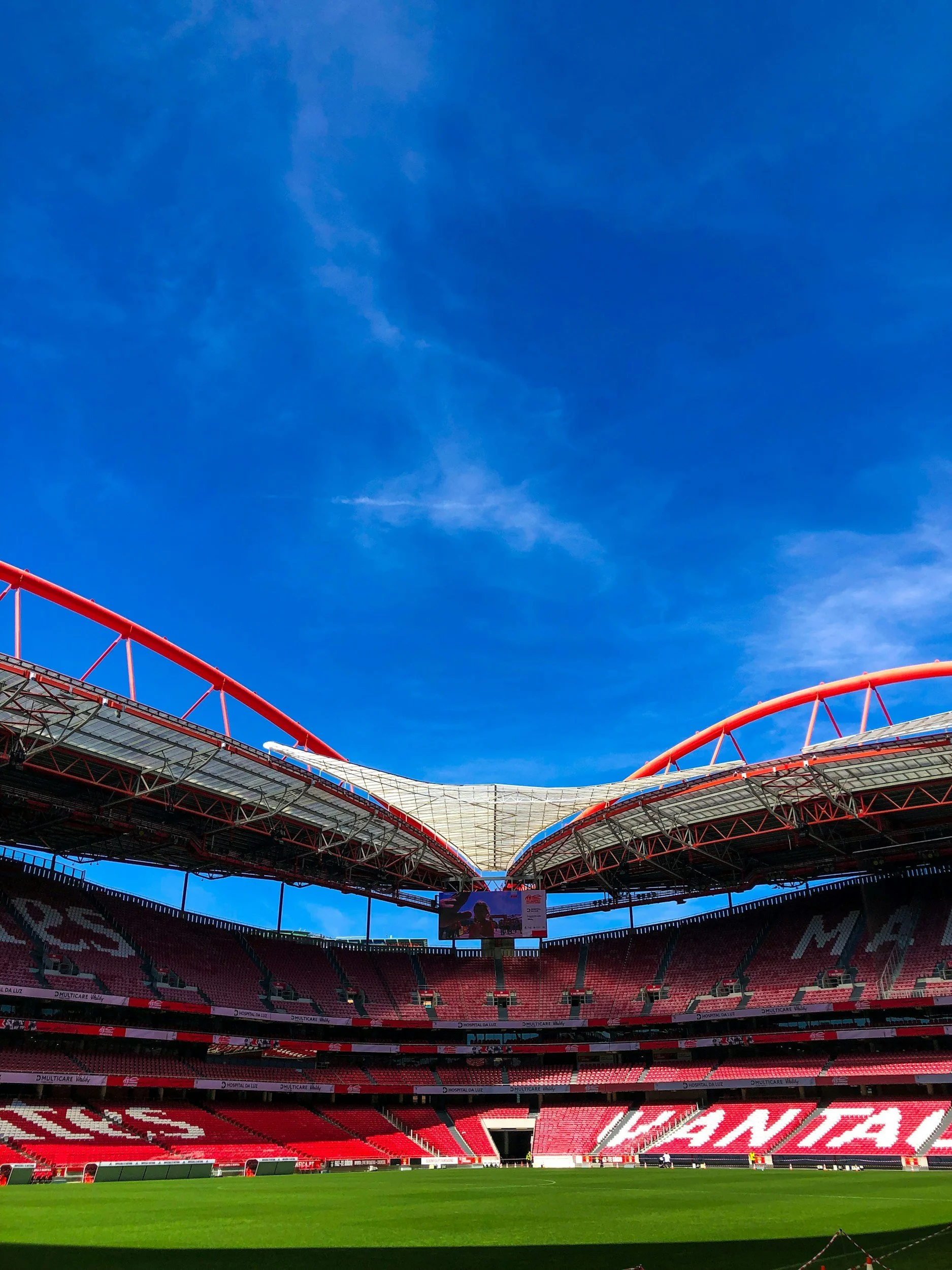 View of a stadium with red seats and a green football field, under a blue sky with some clouds, and the stadium has a partially open roof structure.