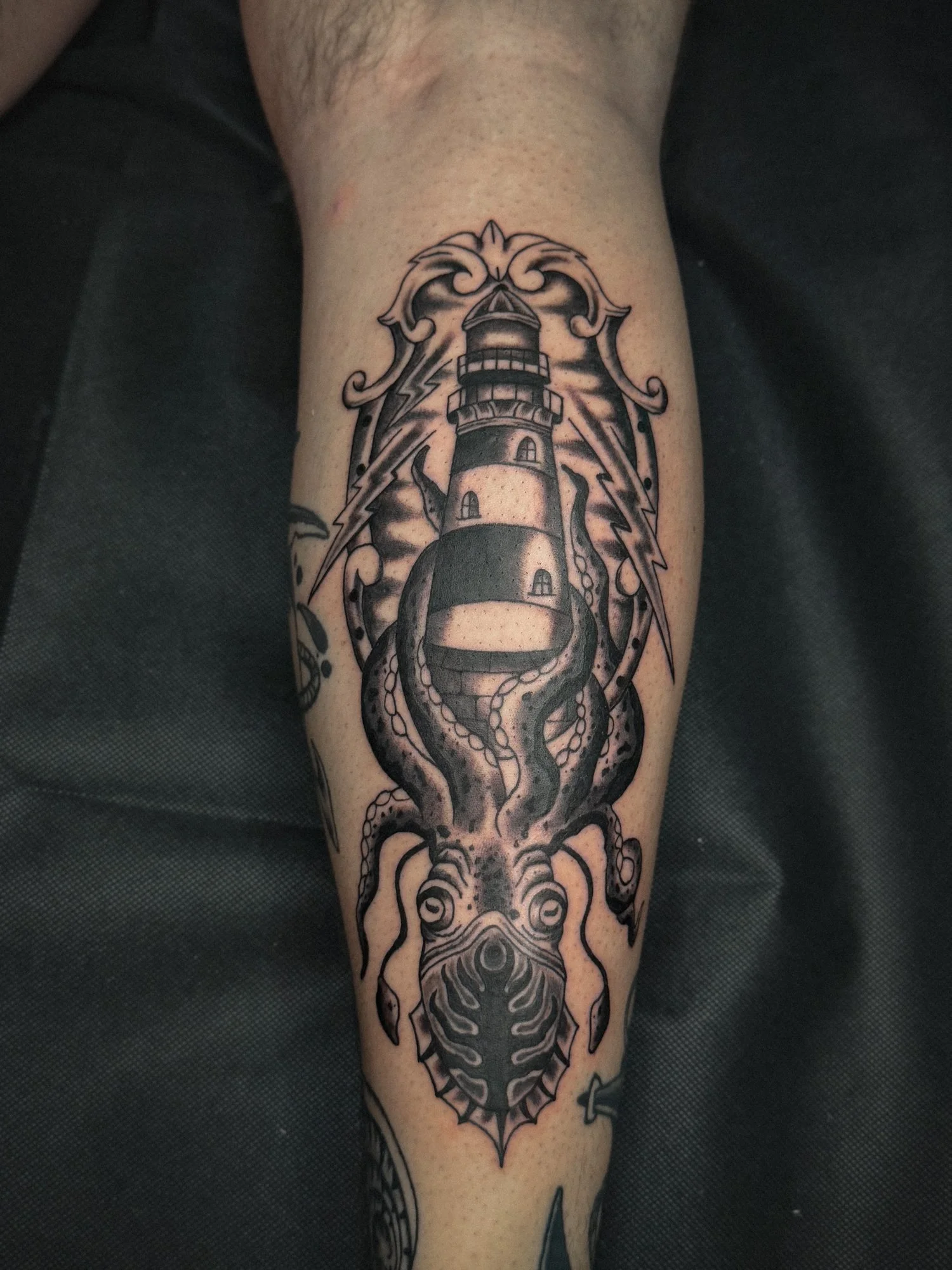 Traditional black and gray tattoo of a squid wrapped around a lighthouse with storm clouds and lightning, framed by an ornamental border.
