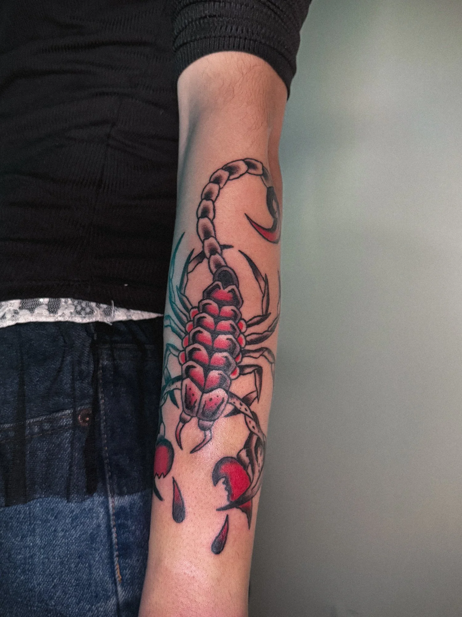 Tattoo of a scorpion on a person's arm with red accents, black outlines, and some green leaves visible around it.