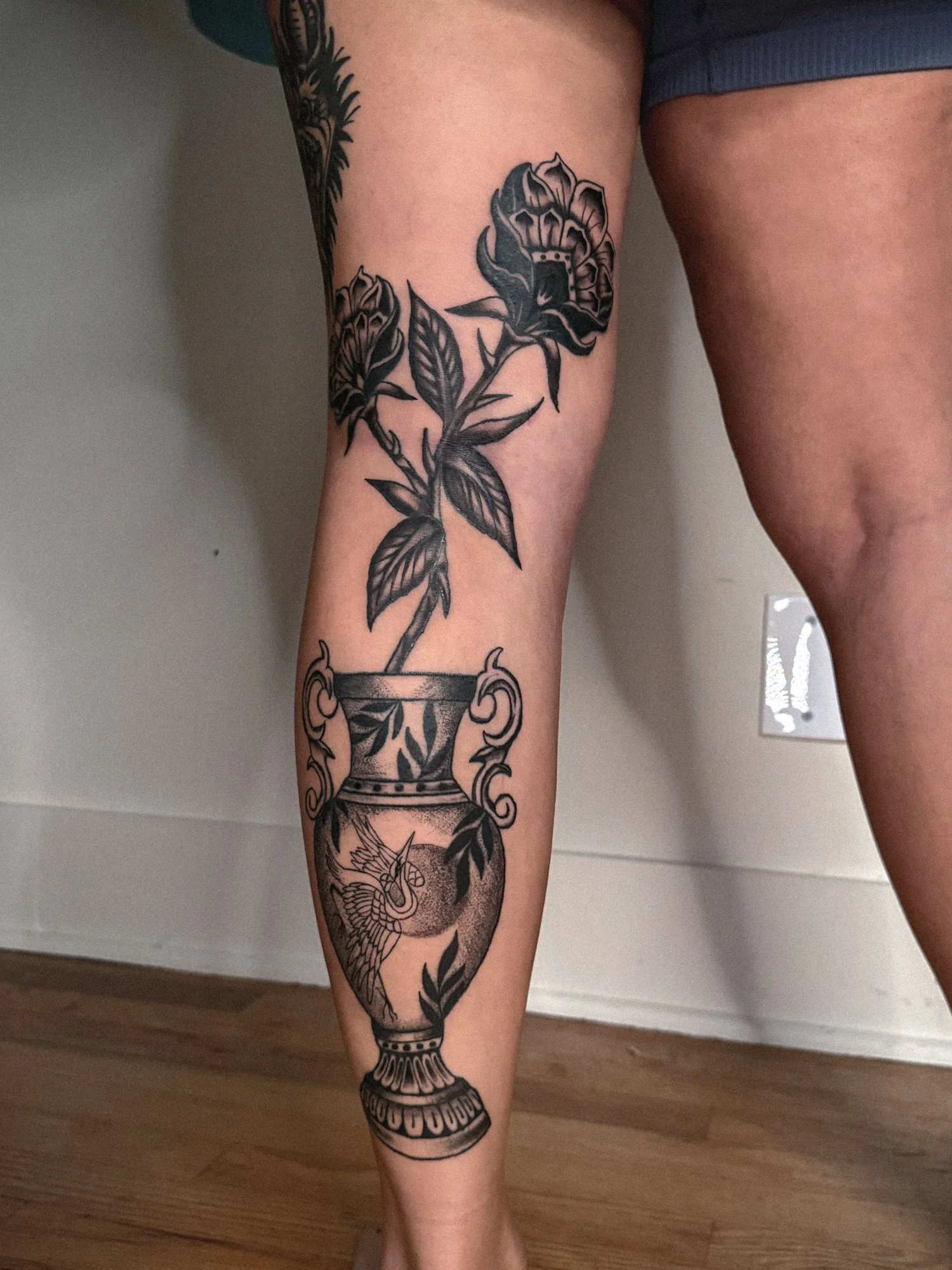 Traditional black and grey flower pot tattoo with two fan flowers, long stems, expressive leaves, and stipple shading in the vase.
