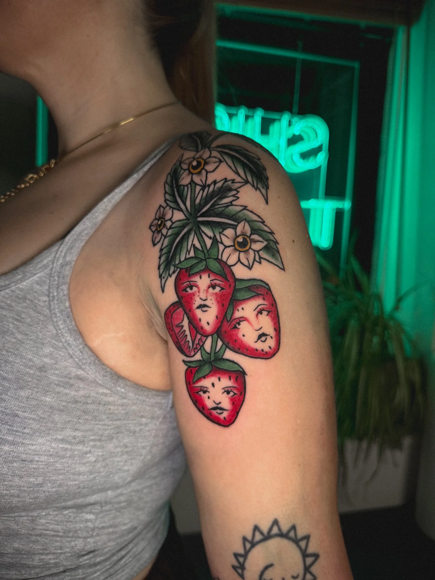 Tattoo of three strawberries with faces, leaves, and flowers on person's upper arm and shoulder.