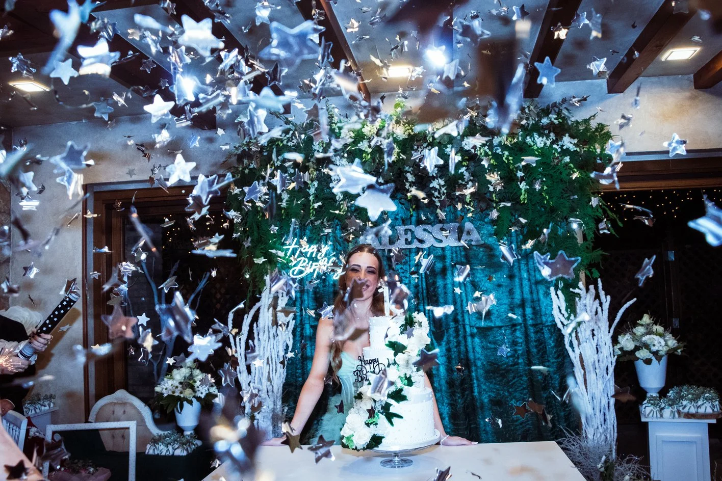A woman at her birthday celebration, standing behind a tiered cake surrounded by floral and decorative elements, with silver star-shaped confetti falling around her.