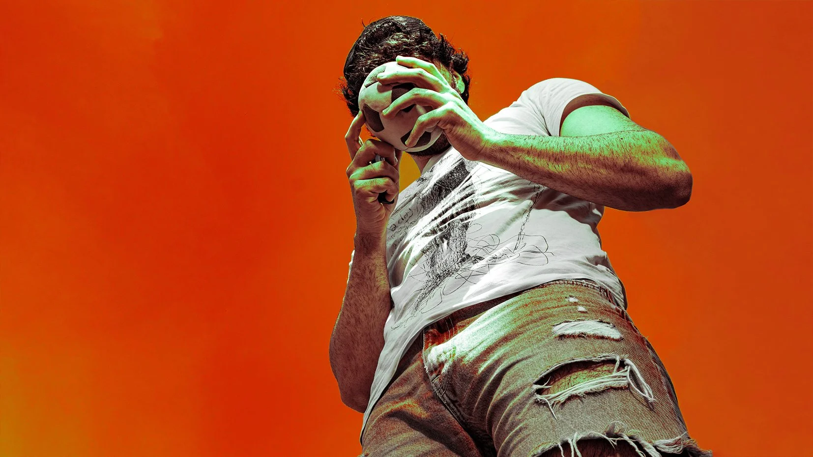 A young man holding a soccer ball to his face, standing against an orange background. He is wearing a white graphic t-shirt and ripped shorts.
