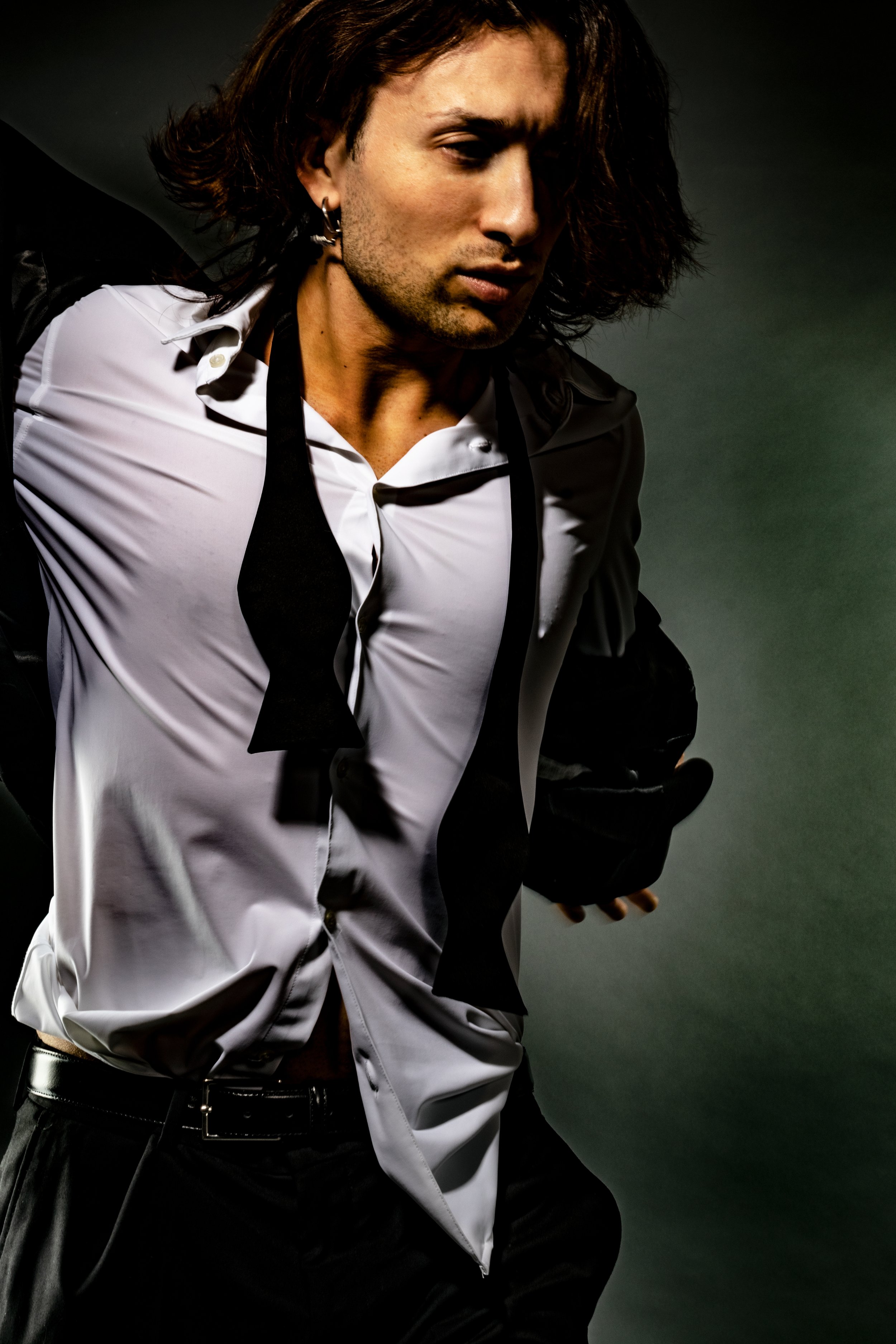 A person with long, wavy brown hair, wearing earrings and a black and white shirt with a black tie, standing against a dark background.