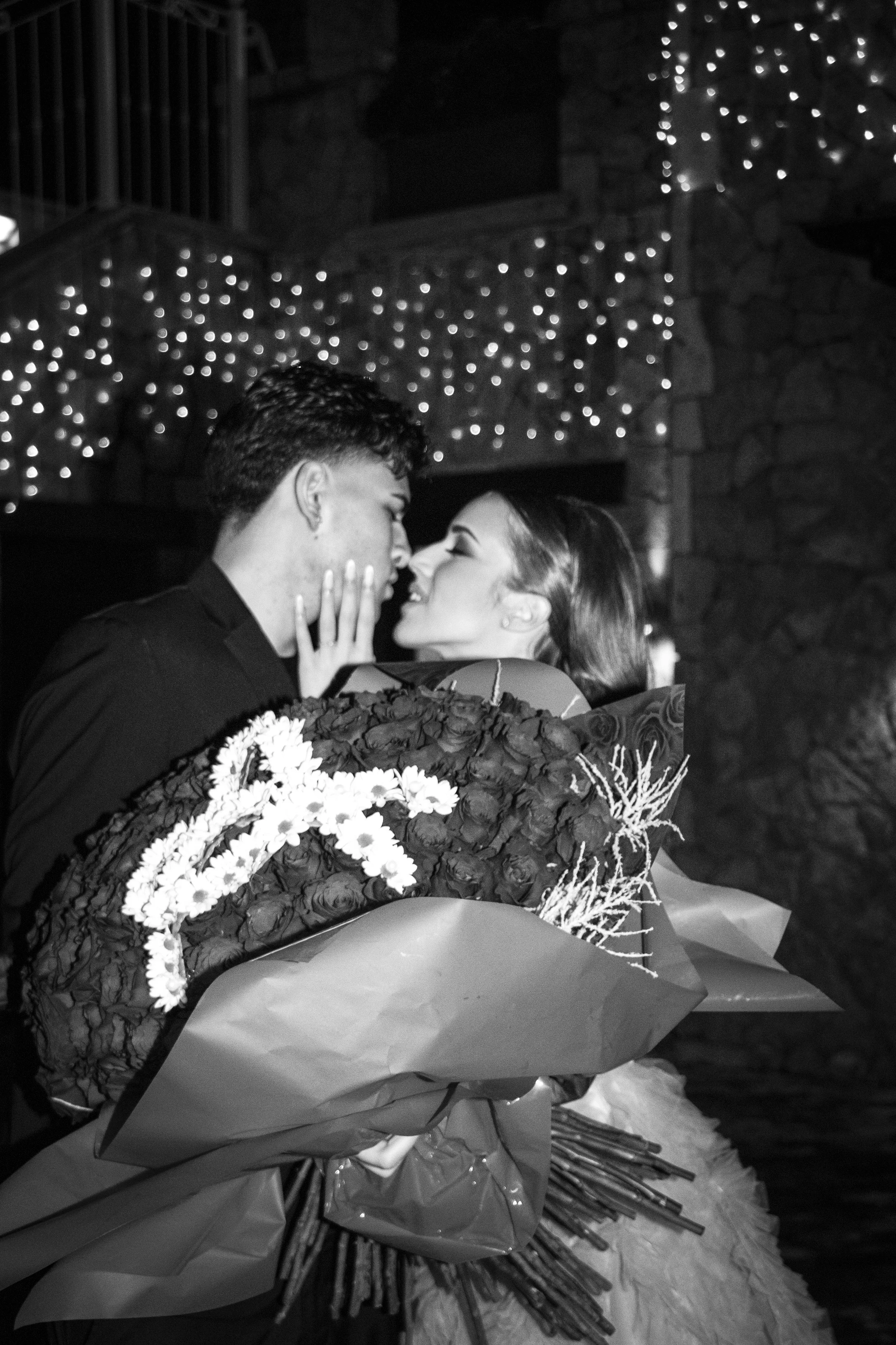 A couple sharing a romantic moment at night, with the woman holding a large bouquet of flowers, in front of decorative string lights.