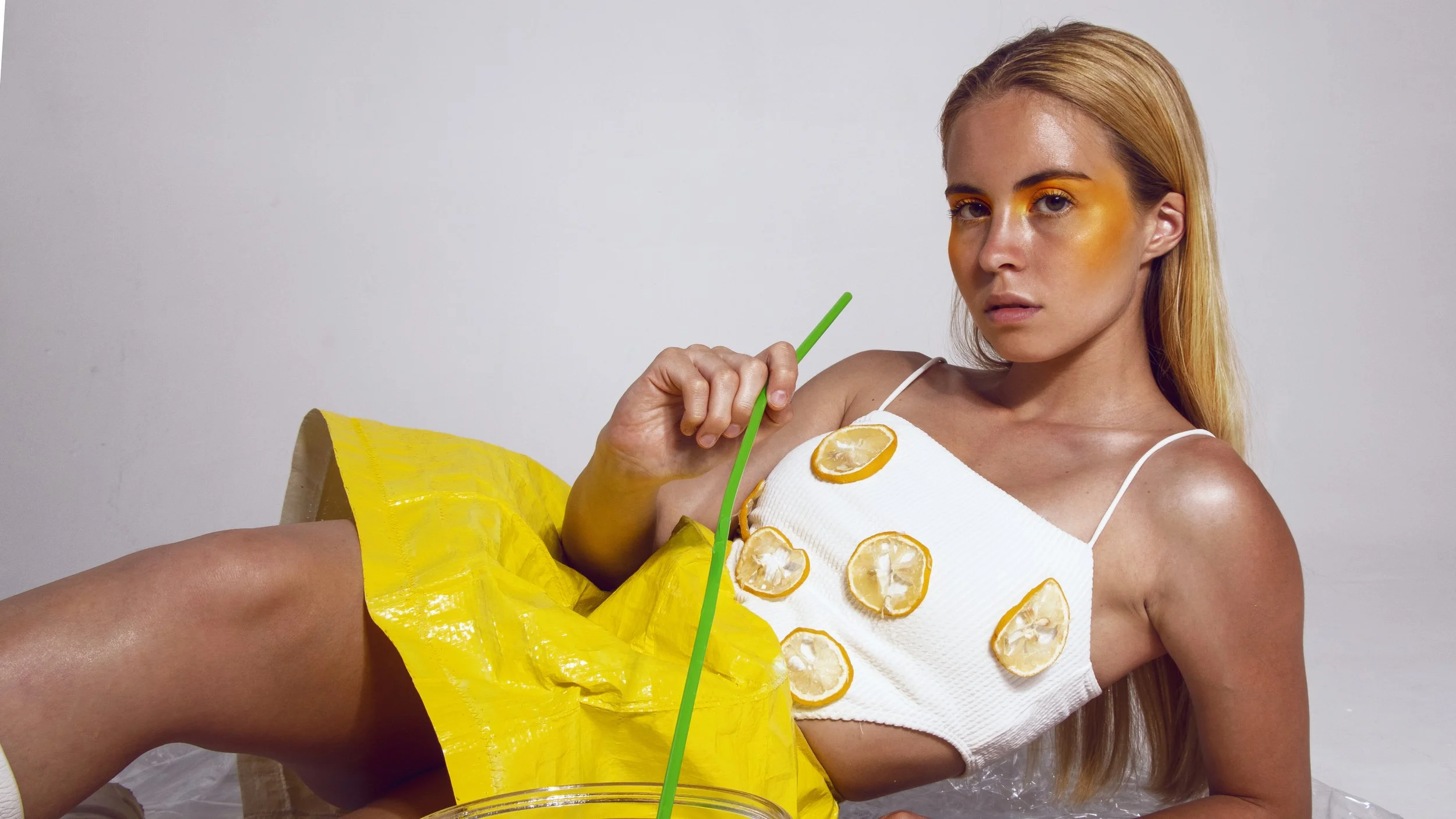 A woman with long blonde hair is lying on a white surface, wearing a white top decorated with lemon slices. She has orange eyeshadow and holds a green straw.