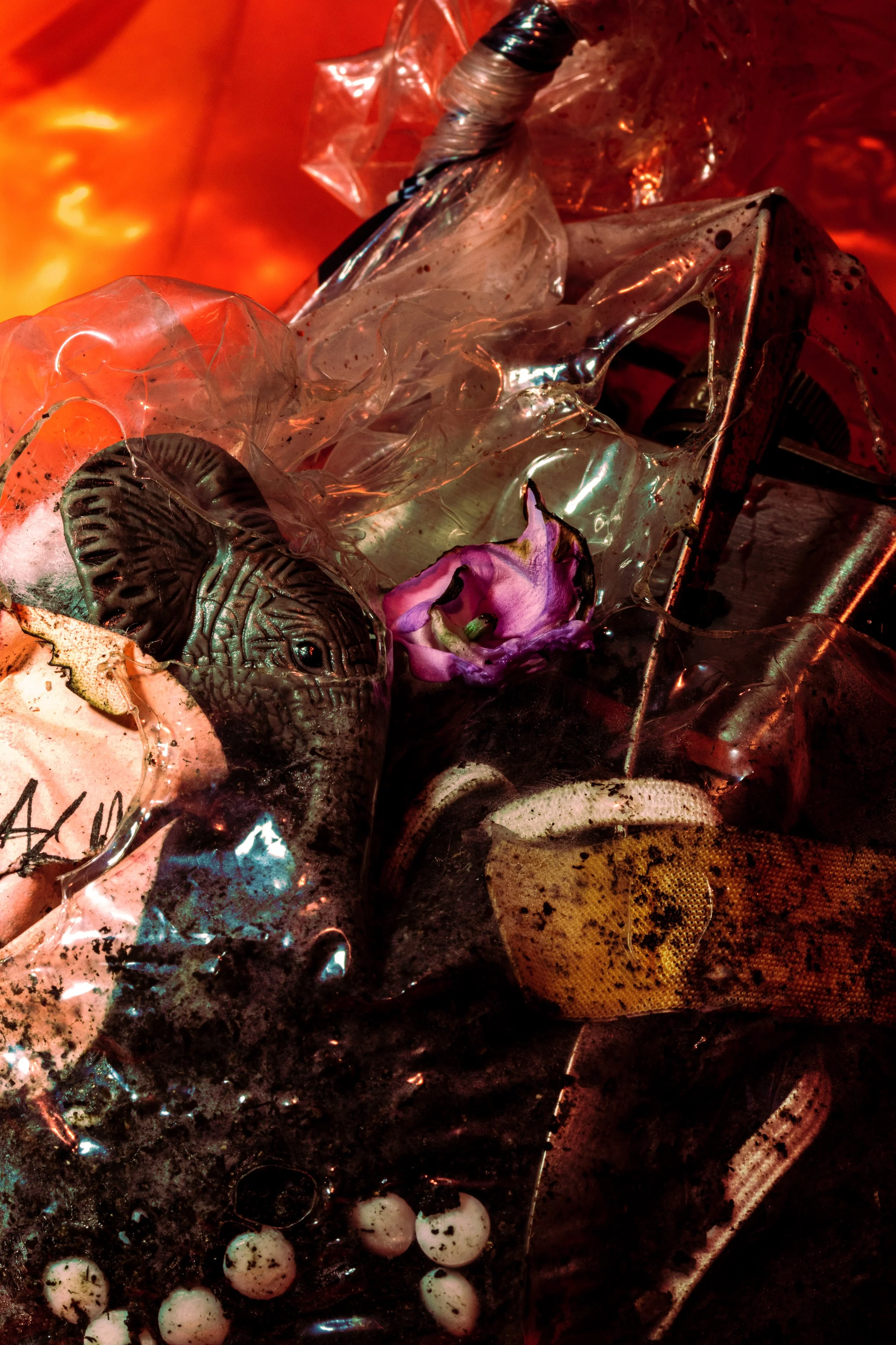 Close-up of trash including a black plastic bag, used plastic wrappers, white plastic balls with black spots, and various debris against an orange-red background.