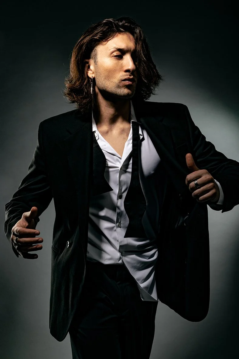 A man with long, wavy hair wearing a black blazer and white shirt, standing against a dark background, posing confidently with his eyes closed, head slightly tilted, and one hand in his pocket.