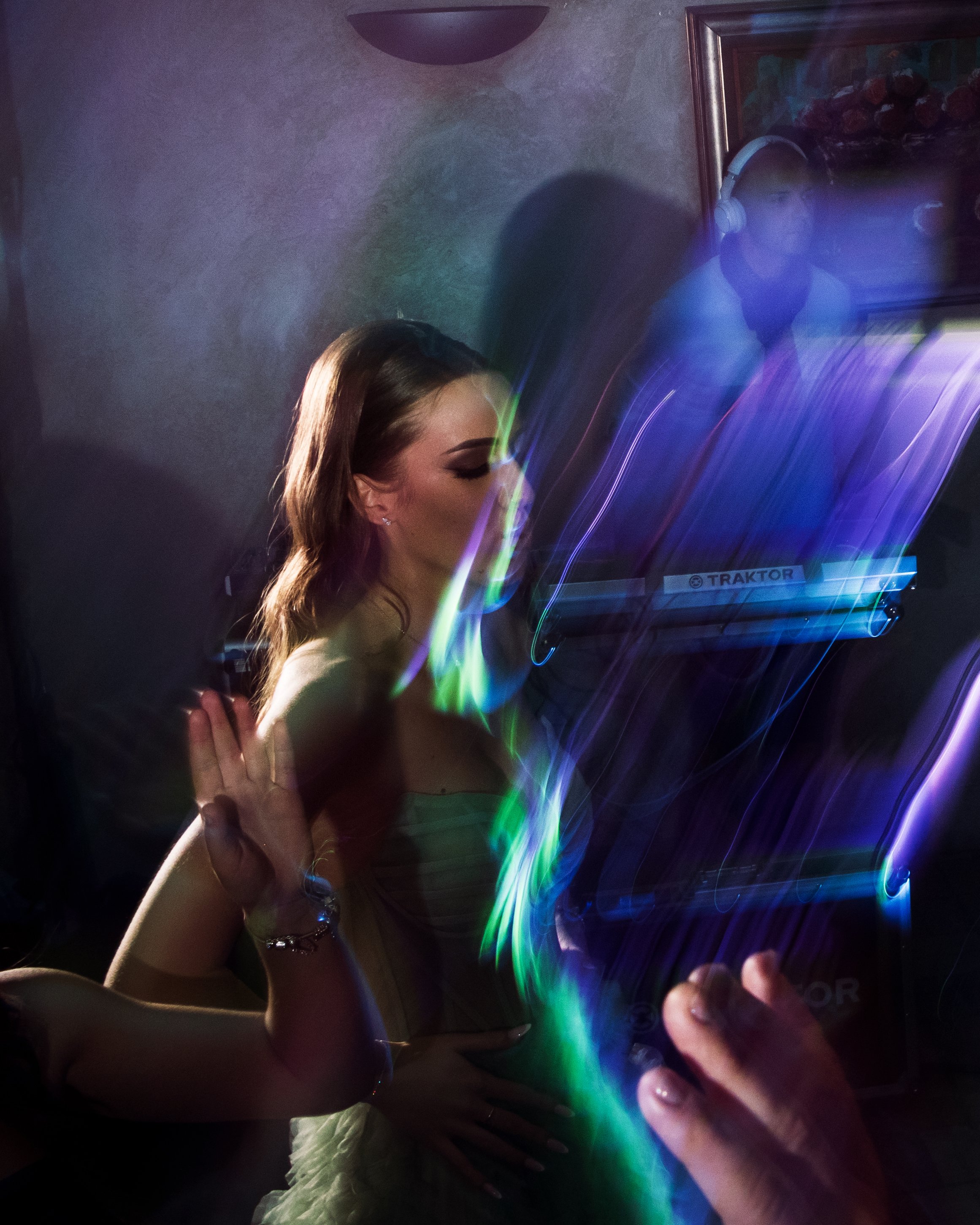 A woman with brown hair, wearing makeup and earrings, is touching a glass or screen that displays a colorful, light streak pattern. Behind the glass, a person with short hair, sunglasses, and headphones is visible. The background features a framed painting of a rocky waterfall scene and a wall with a textured surface.