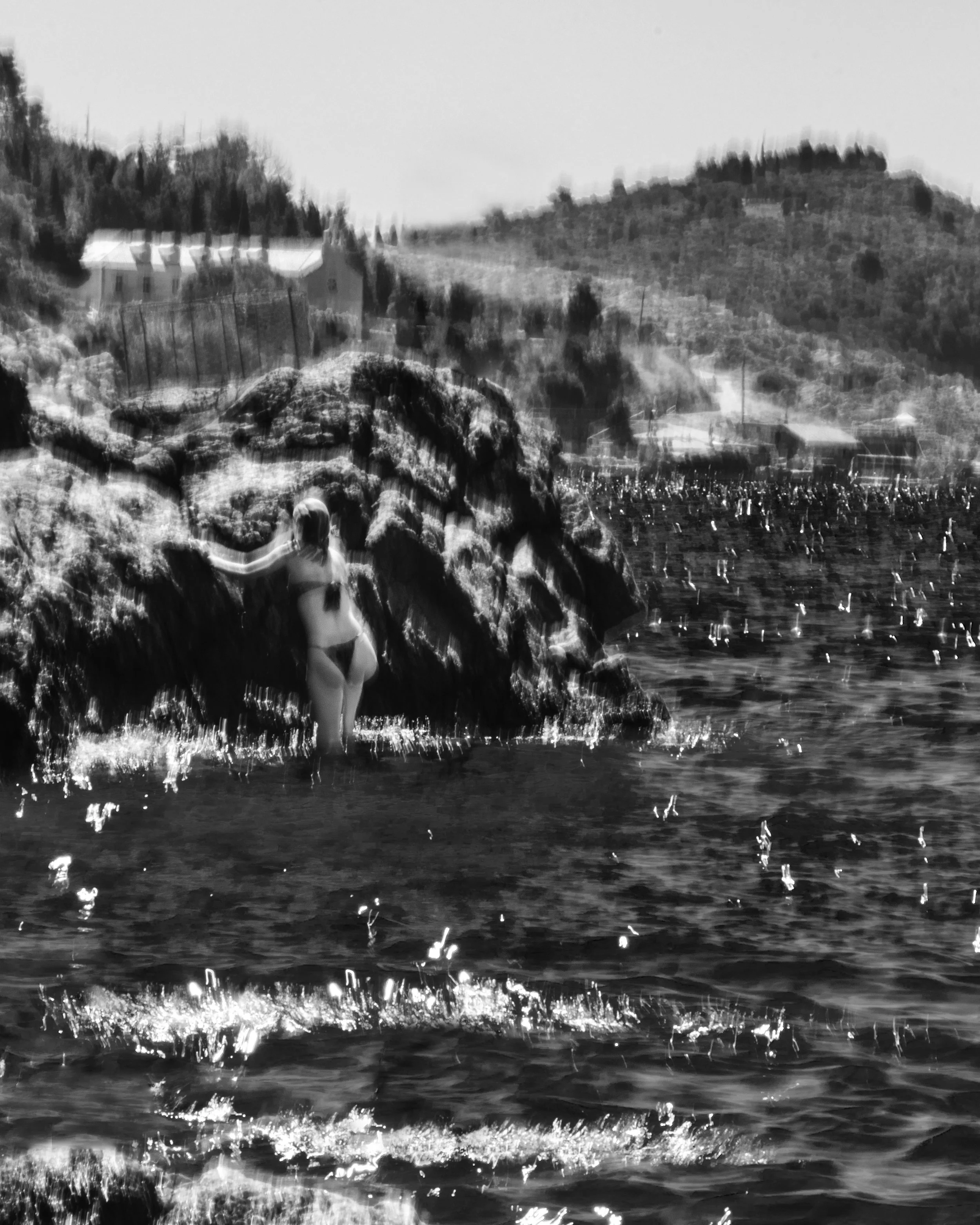 A black and white photo of a person in the water, wearing a swimsuit, near rocks, with a hillside in the background and a building on top of the hill.