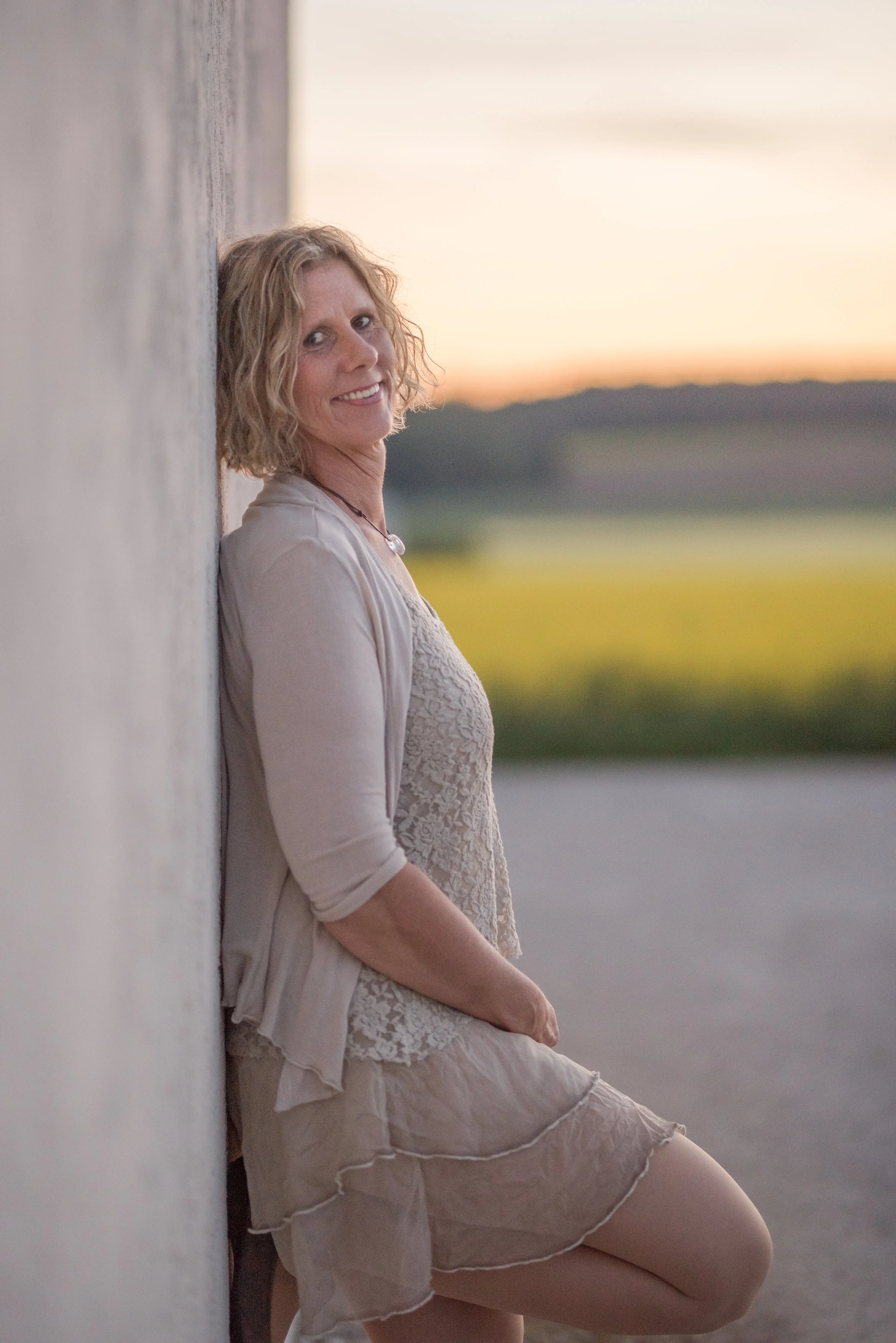 Eine Frau mit lockigem Haar lehnt an einer weißen Wand und trägt ein helles, sommerliches Outfit. Im Hintergrund ist ein Sonnenuntergang mit einer ländlichen Landschaft zu sehen.