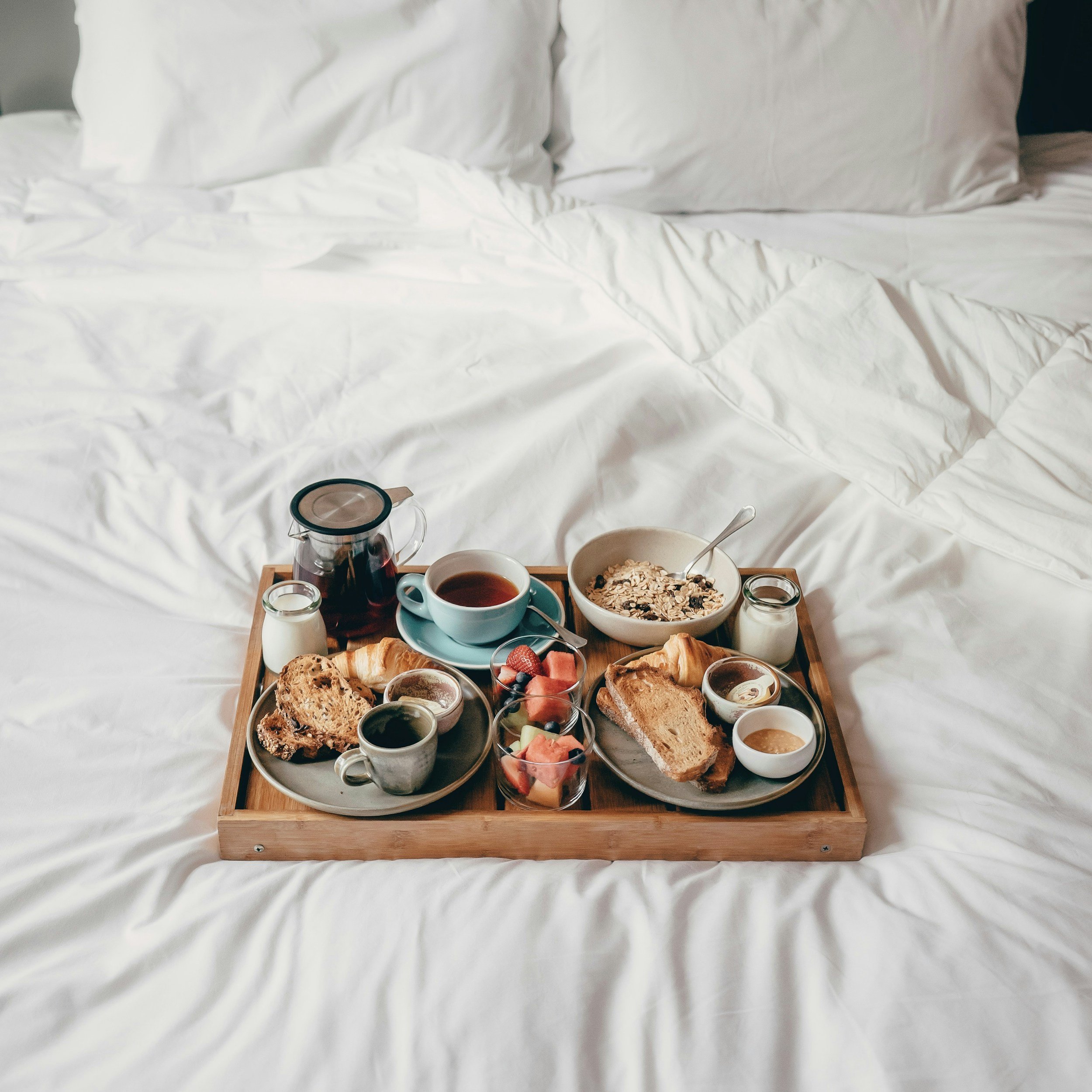 A breakfast tray on a bed with white sheets, holding a variety of breakfast foods including bread, fruit, cereal, coffee, and small jars of condiments.