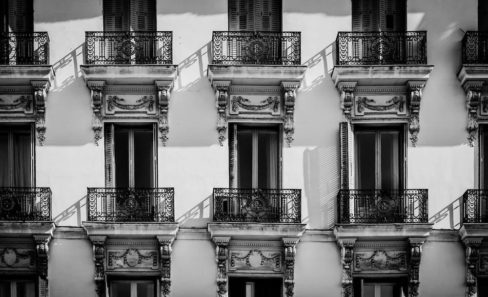 Black and white photo of a building facade with three floors, each featuring balconies with ornate wrought iron railings and decorative architectural details around windows and balconies.