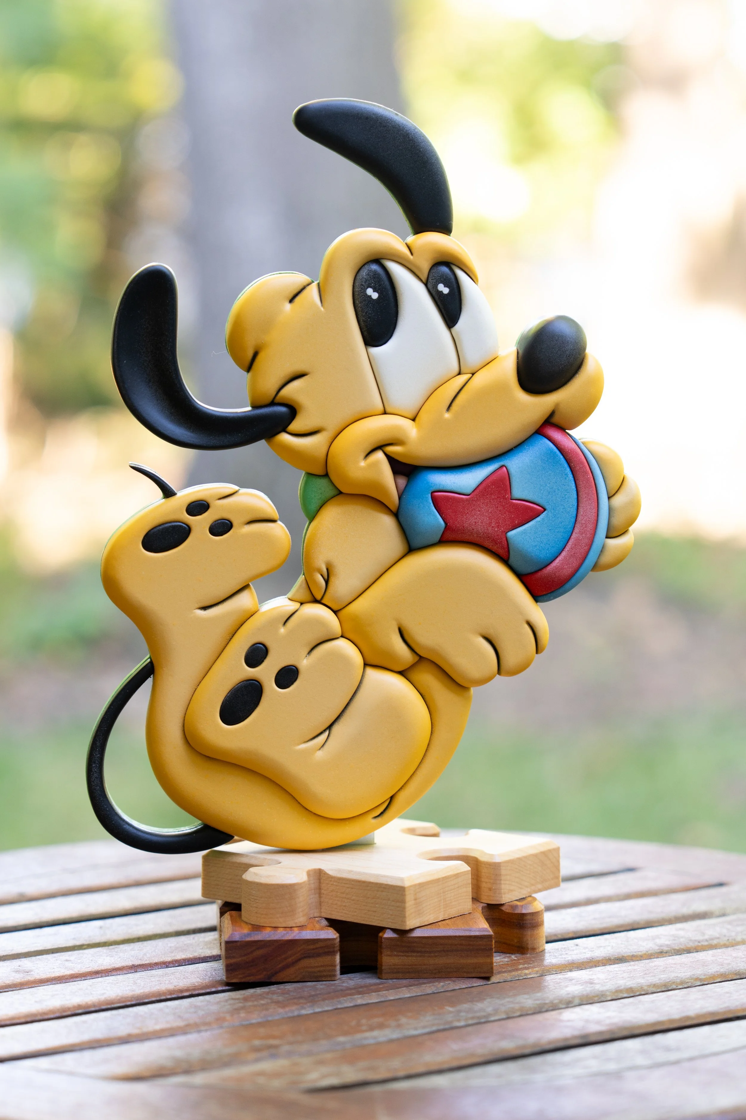 Colorful figurine of Pluto, a  cartoon dog, with a yellow body, black ears, and a red star on its blue chest, standing on a wooden base outdoors with a blurred background of trees.